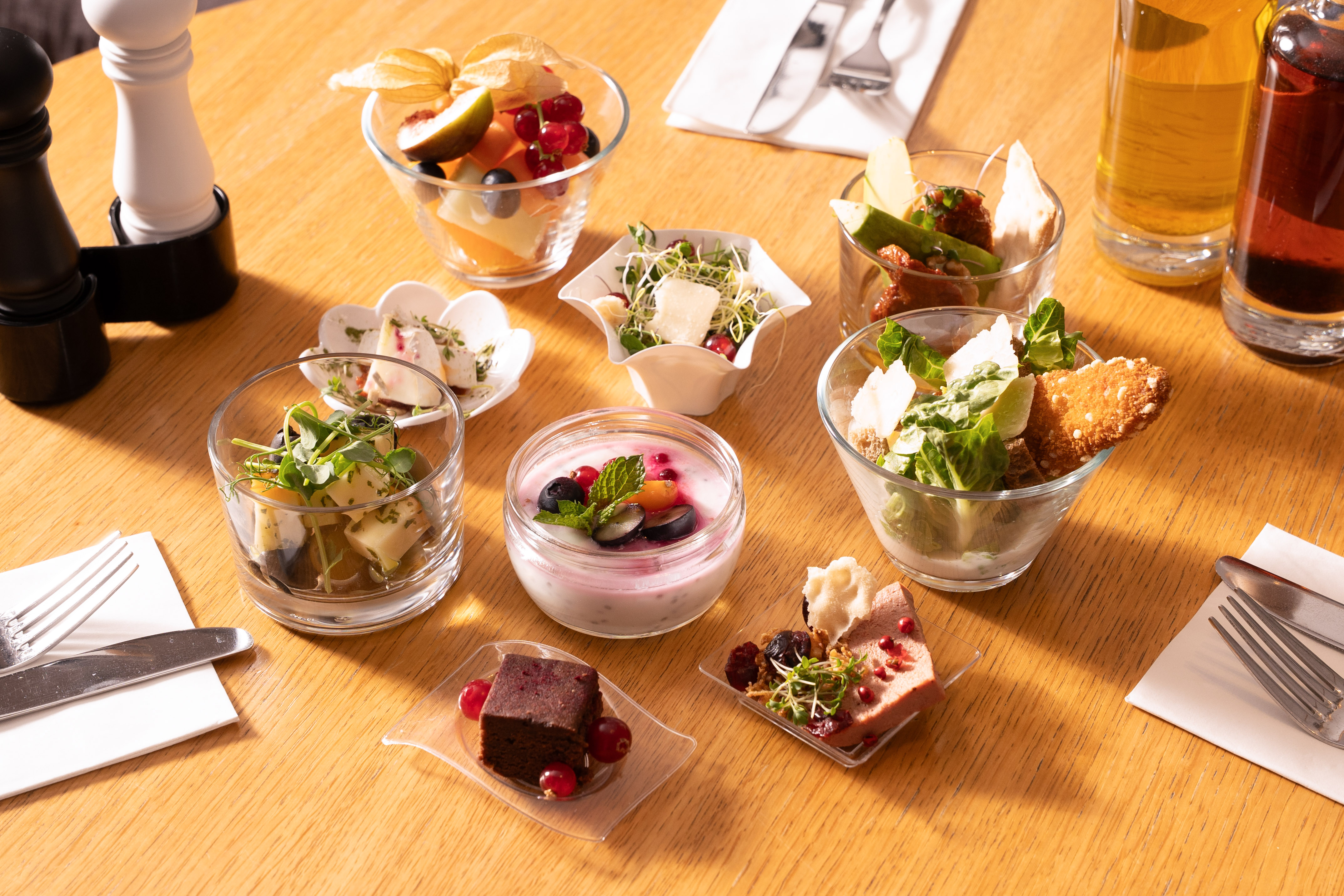 Varied mini food buffet with salads, fruit, yogurt, and desserts on a wood table.