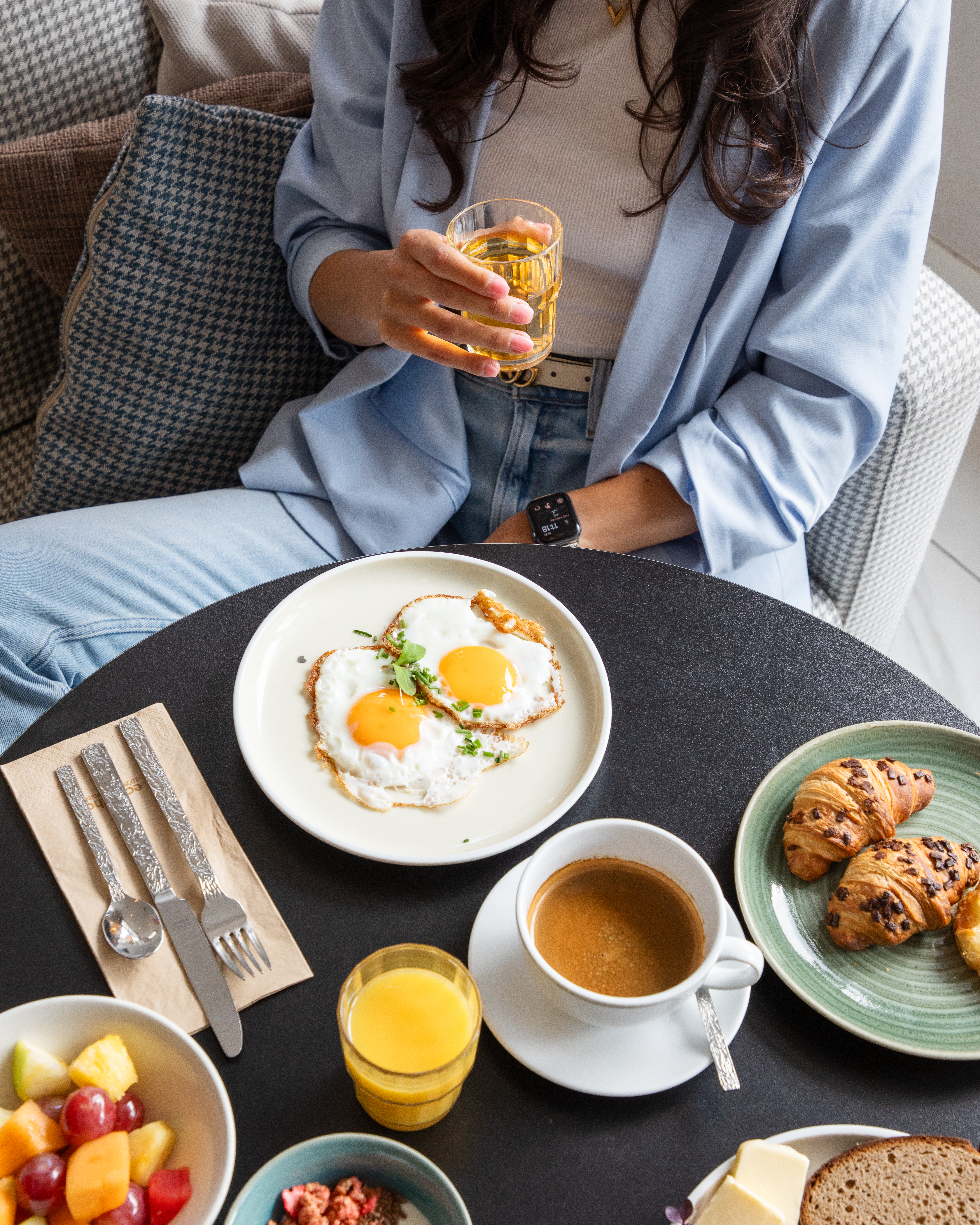 Mujer desayunando con huevos fritos, café, cruasanes, fruta y zumo.