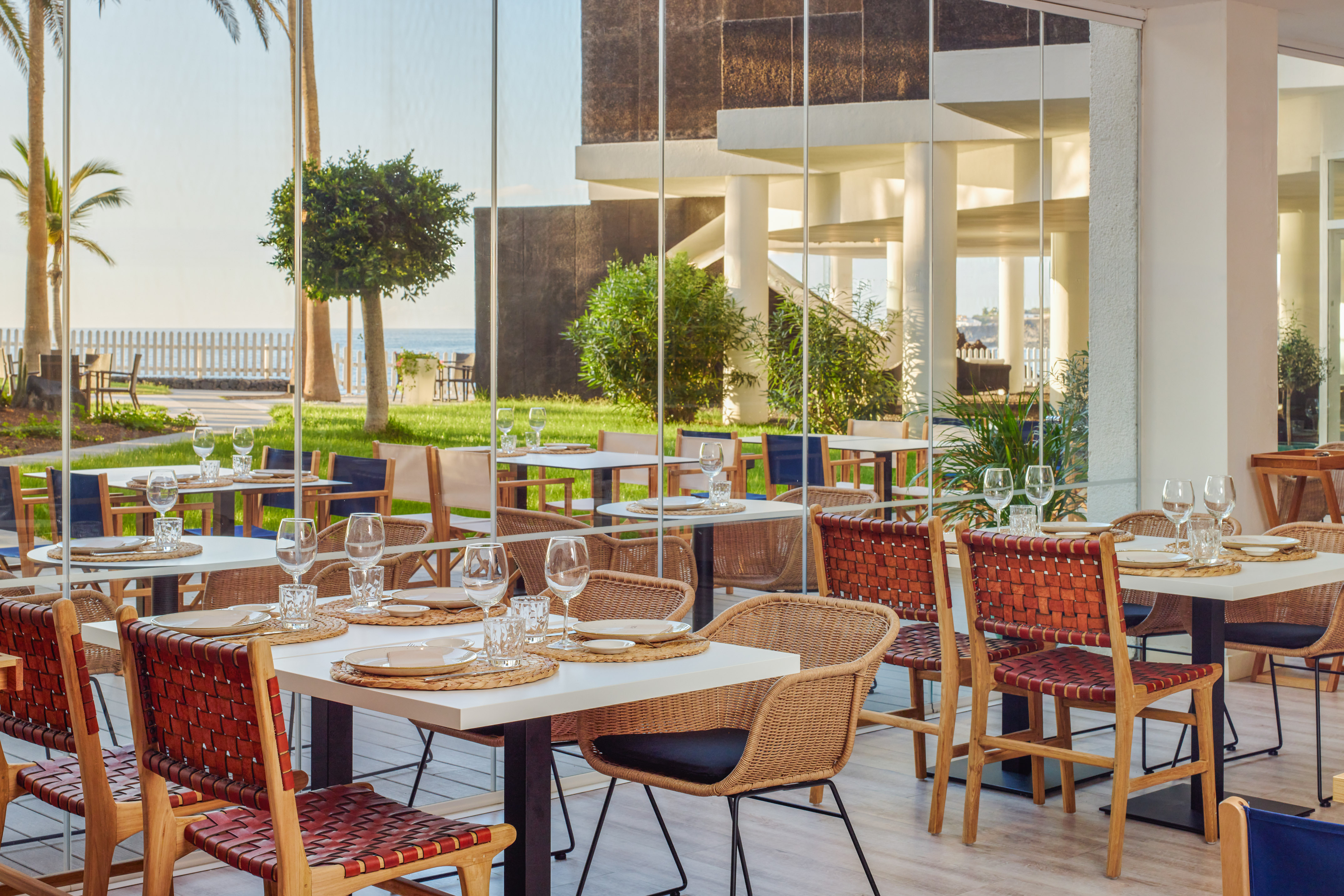 Modern coastal restaurant terrace with ocean view, wicker and leather chairs, set tables.
