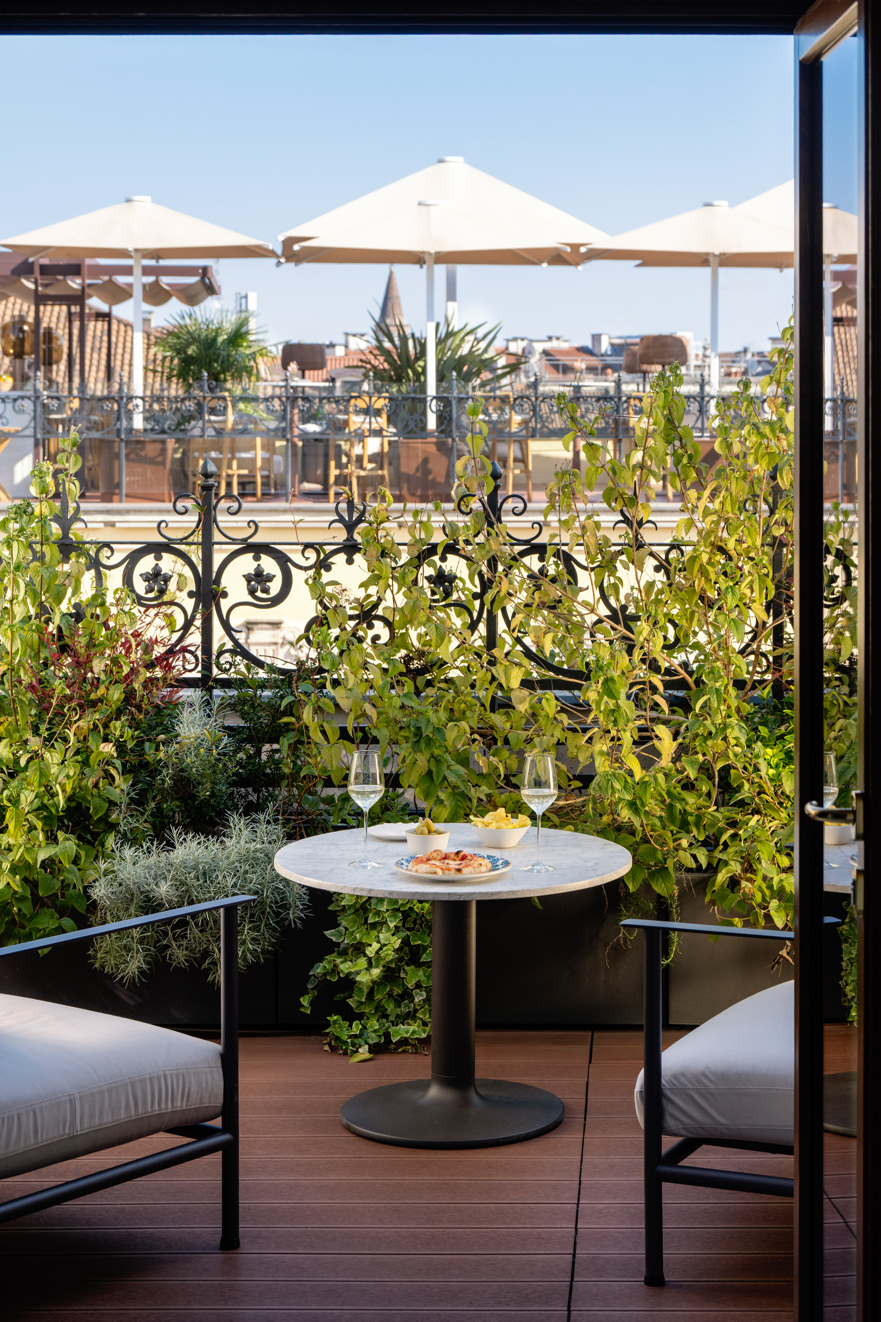 Rooftop terrace with marble table, wine, snacks, and lush plants, city view.
