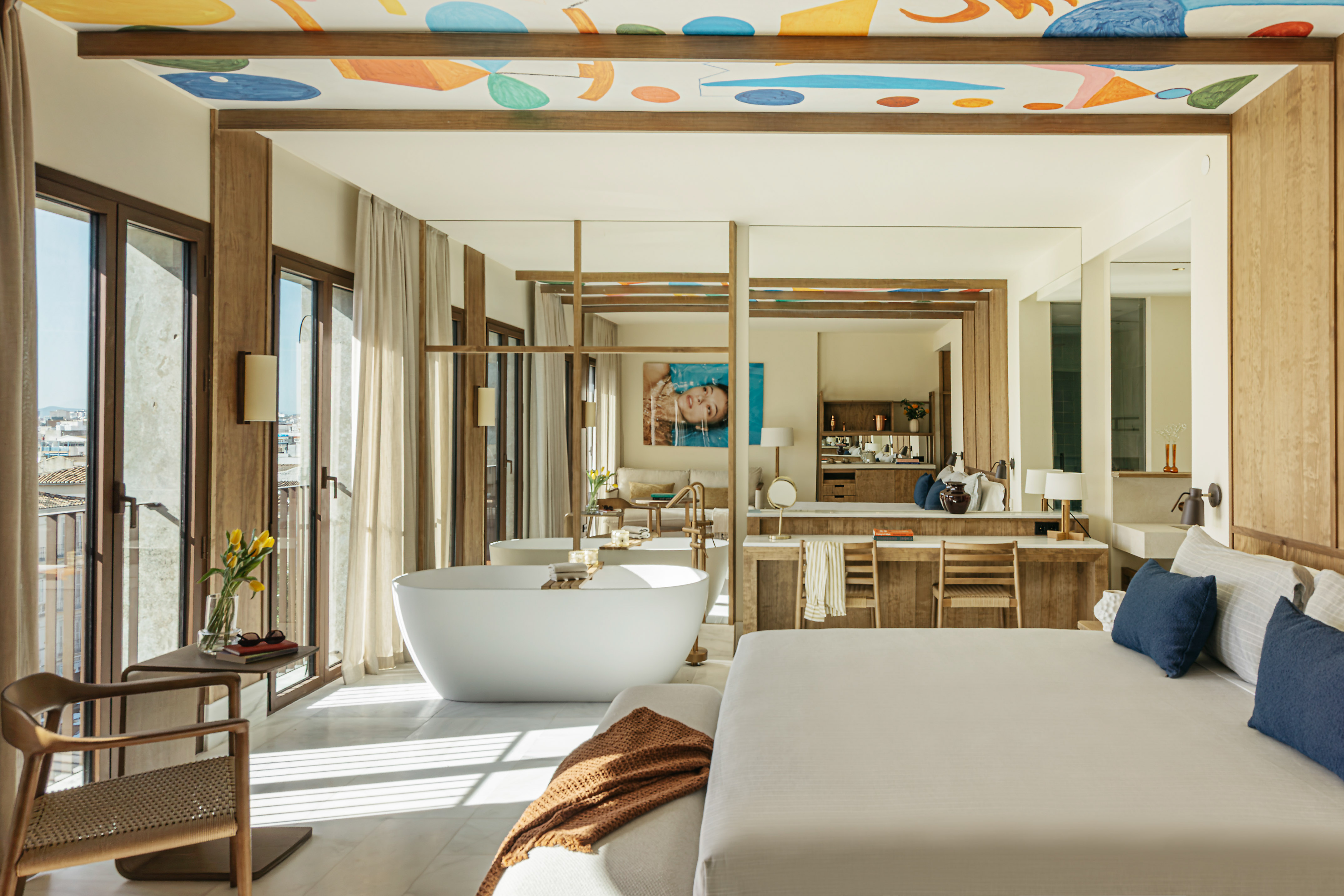 Luxury hotel suite with freestanding tub, modern bed, and colorful ceiling art.