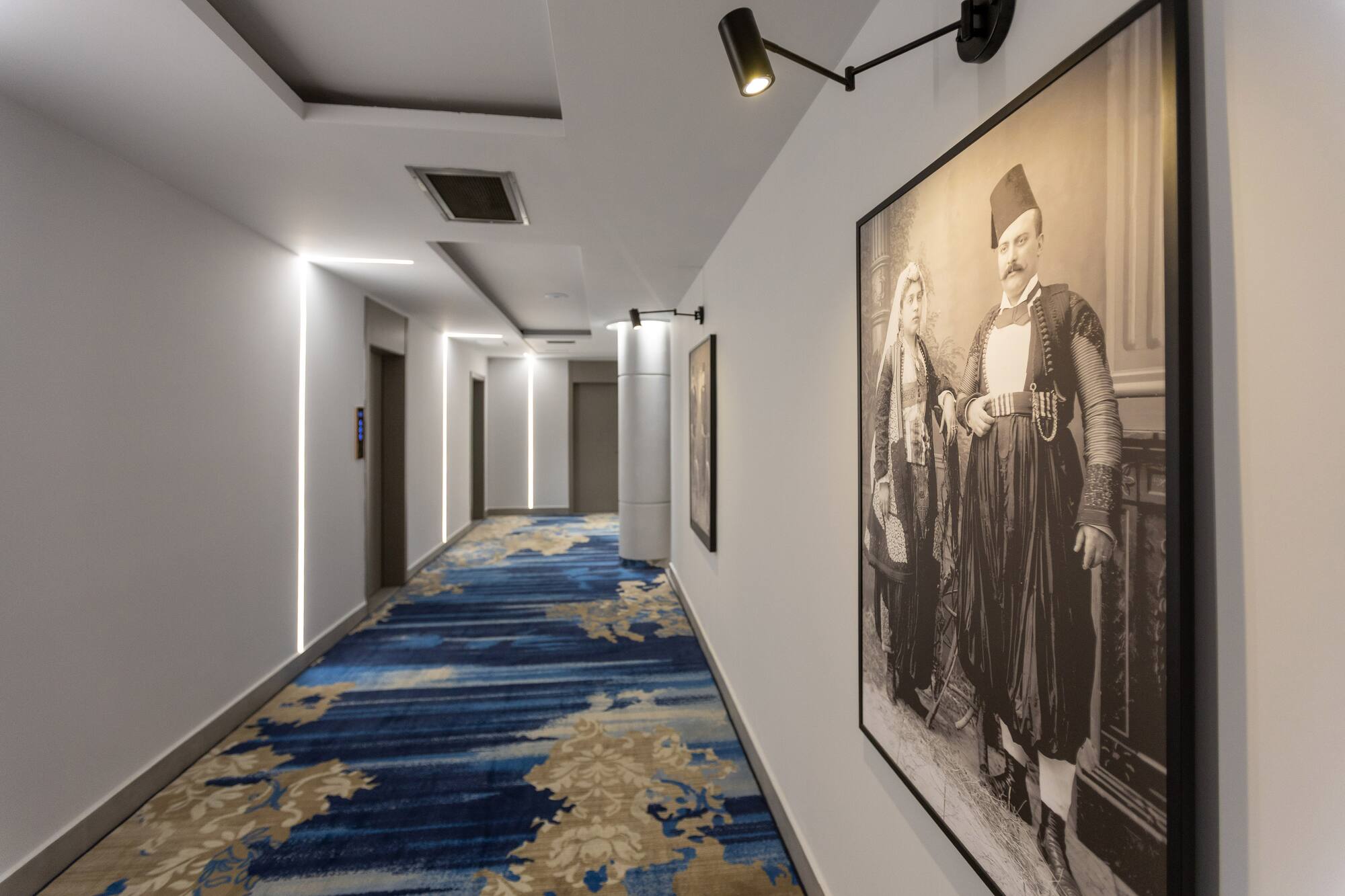 Modern hotel hallway with blue patterned carpet, accent lighting, and traditional portrait.