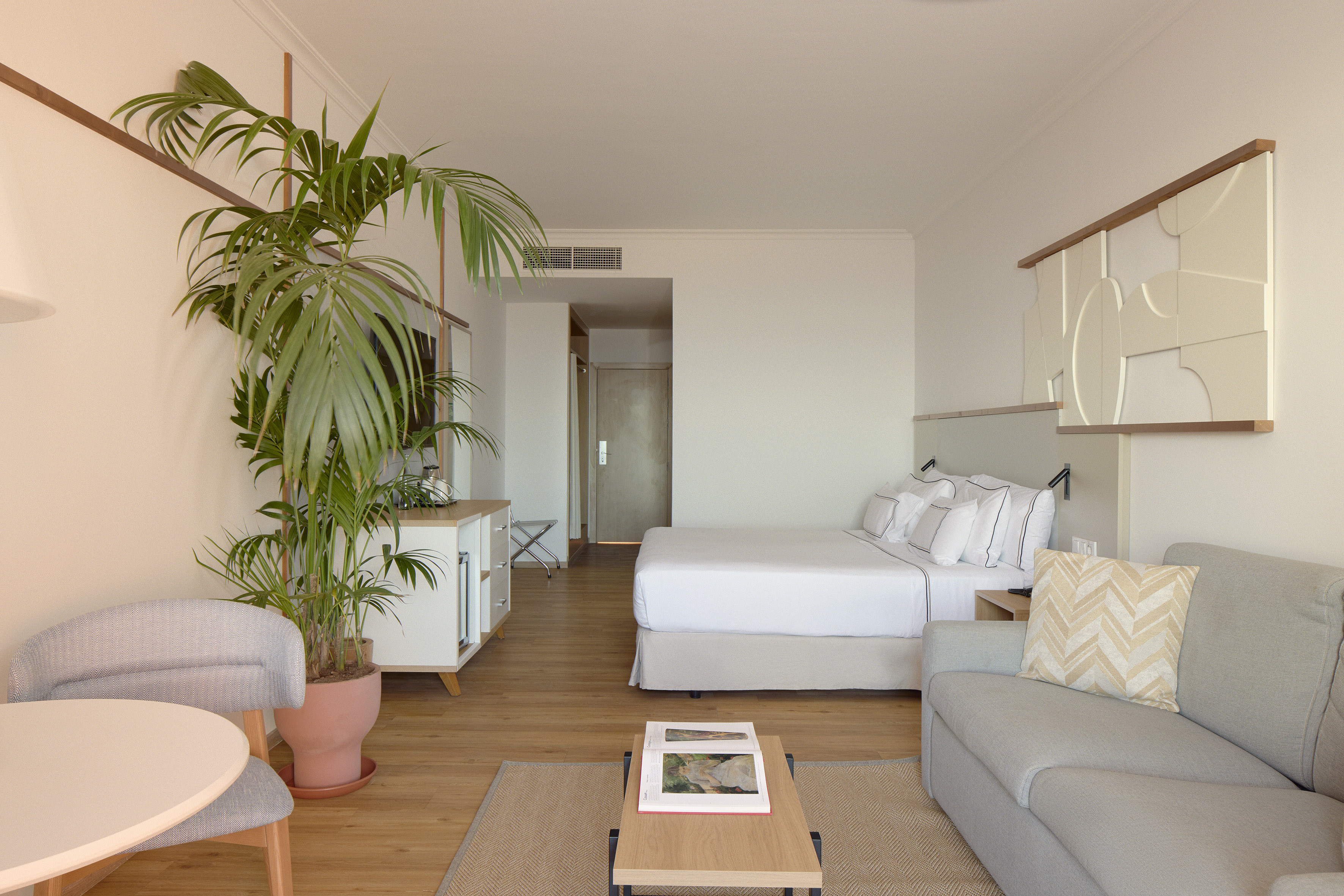 Modern hotel suite with bed, sofa, dining area, and large palm plant.