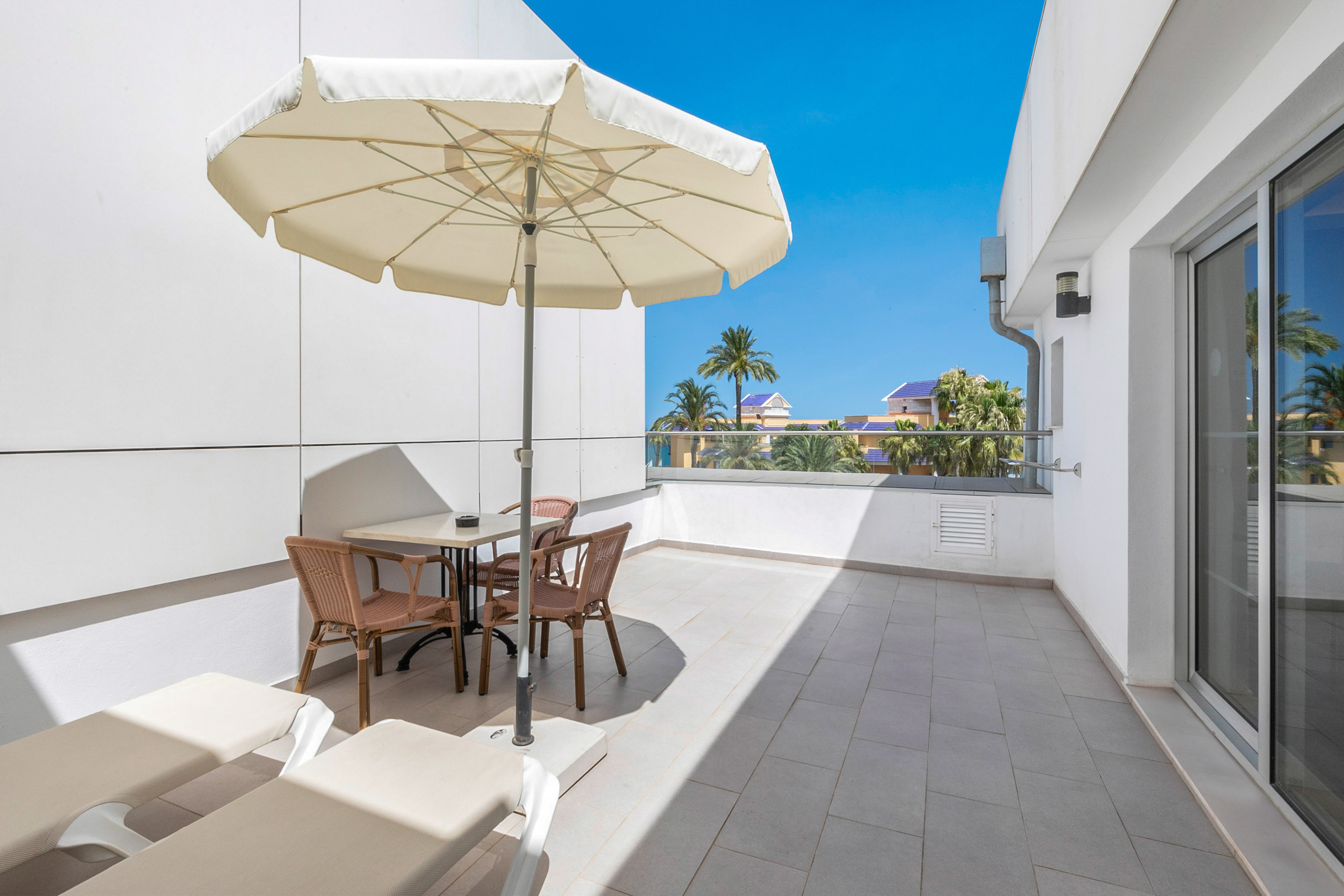 Sunny balcony with patio umbrella, dining set, lounge chairs, and palm trees.