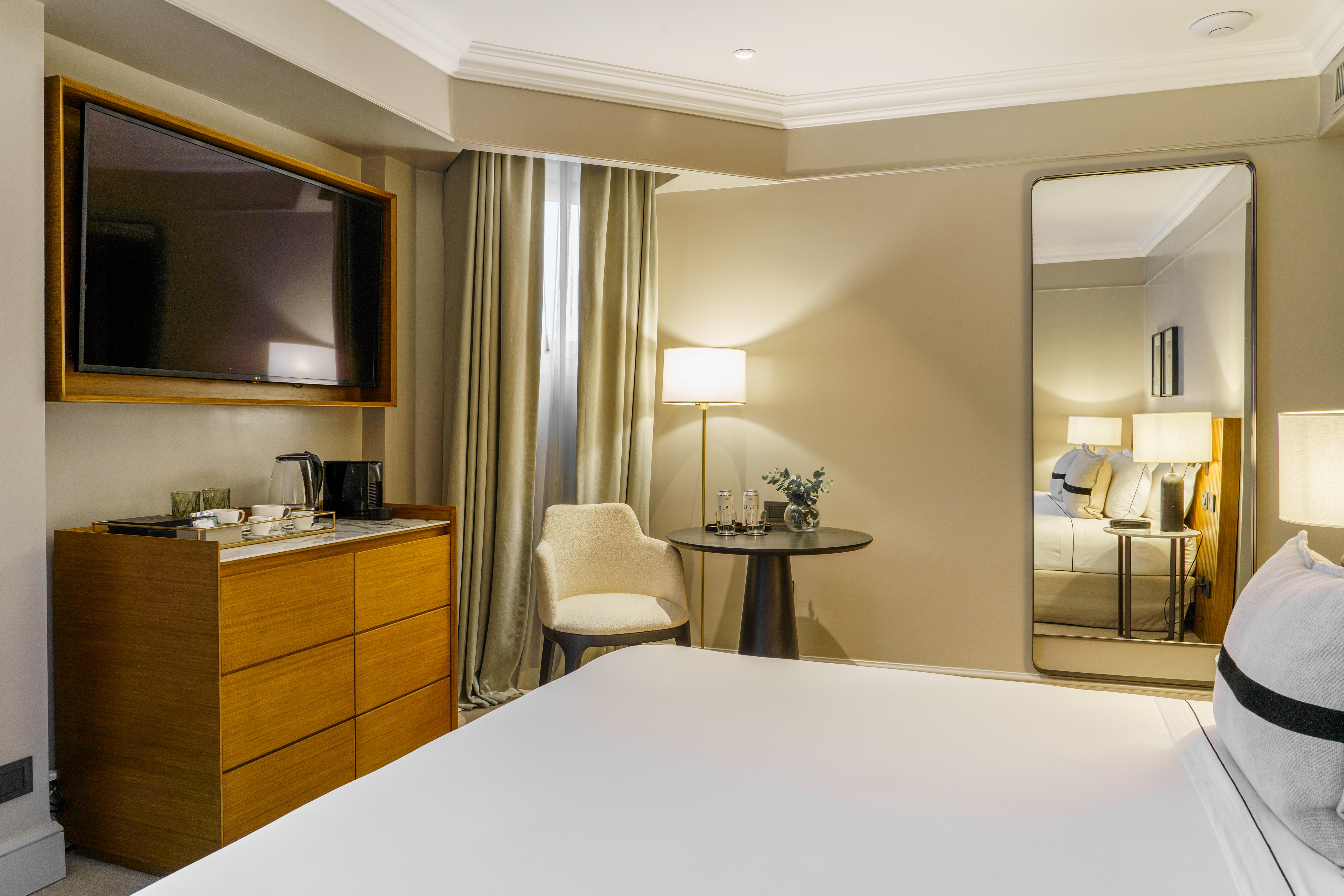 Modern hotel bedroom with TV, white bed, coffee station, and full-length mirror.