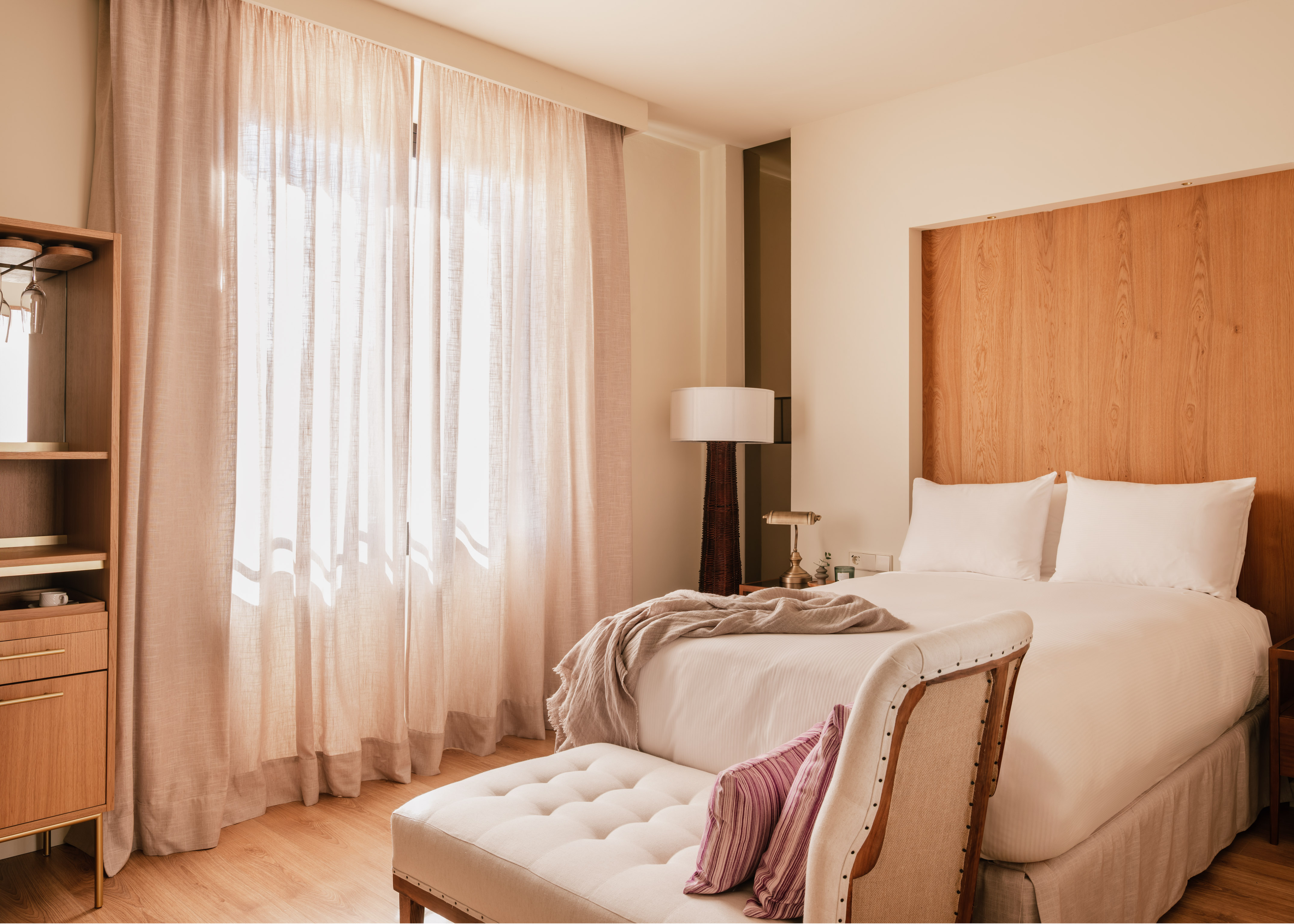 Elegant hotel bedroom with wooden headboard, white bed, and sheer curtains.