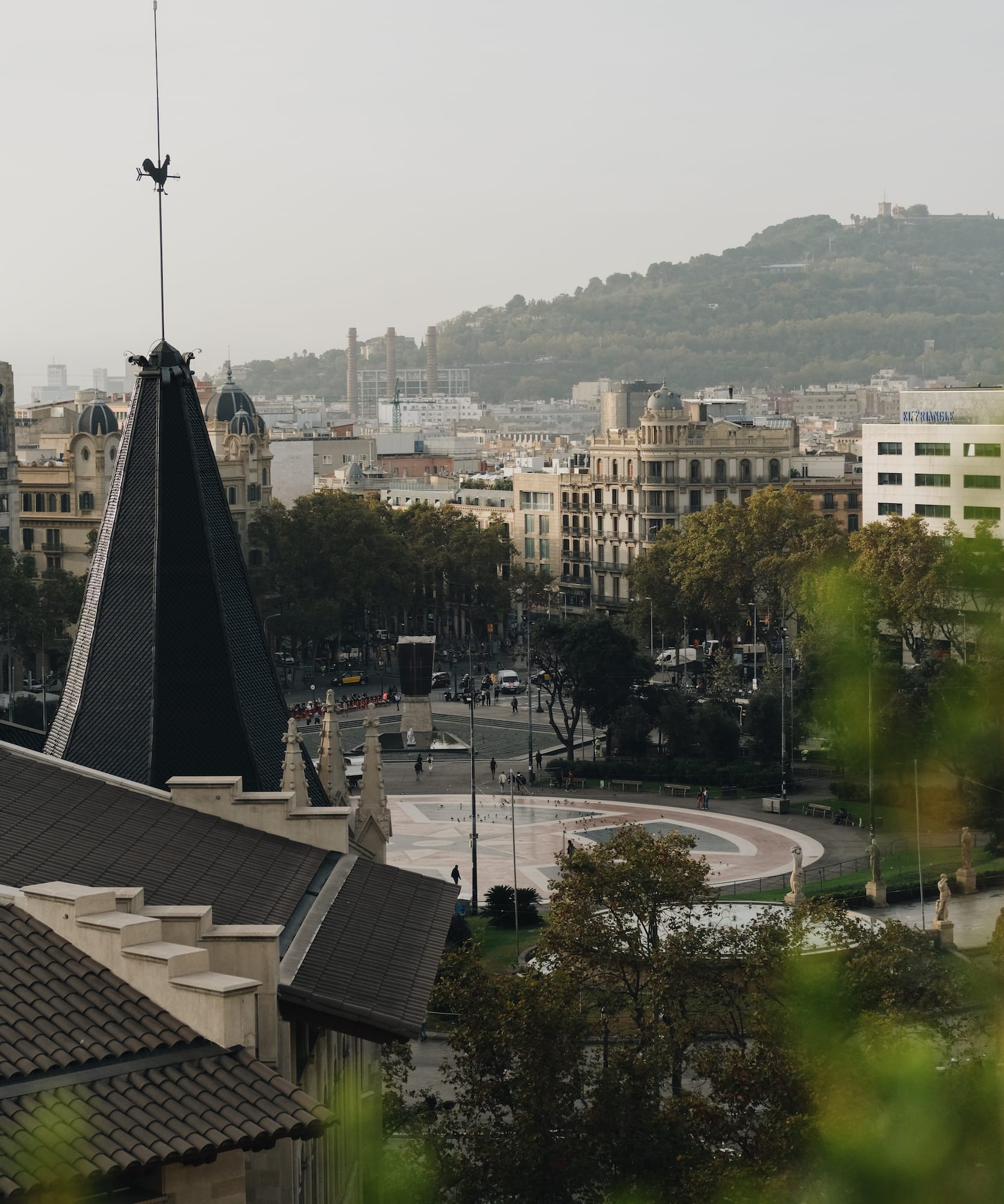 Panorama de Plaça Catalunya, Barcelona: tejado con veleta, edificios, montaña.