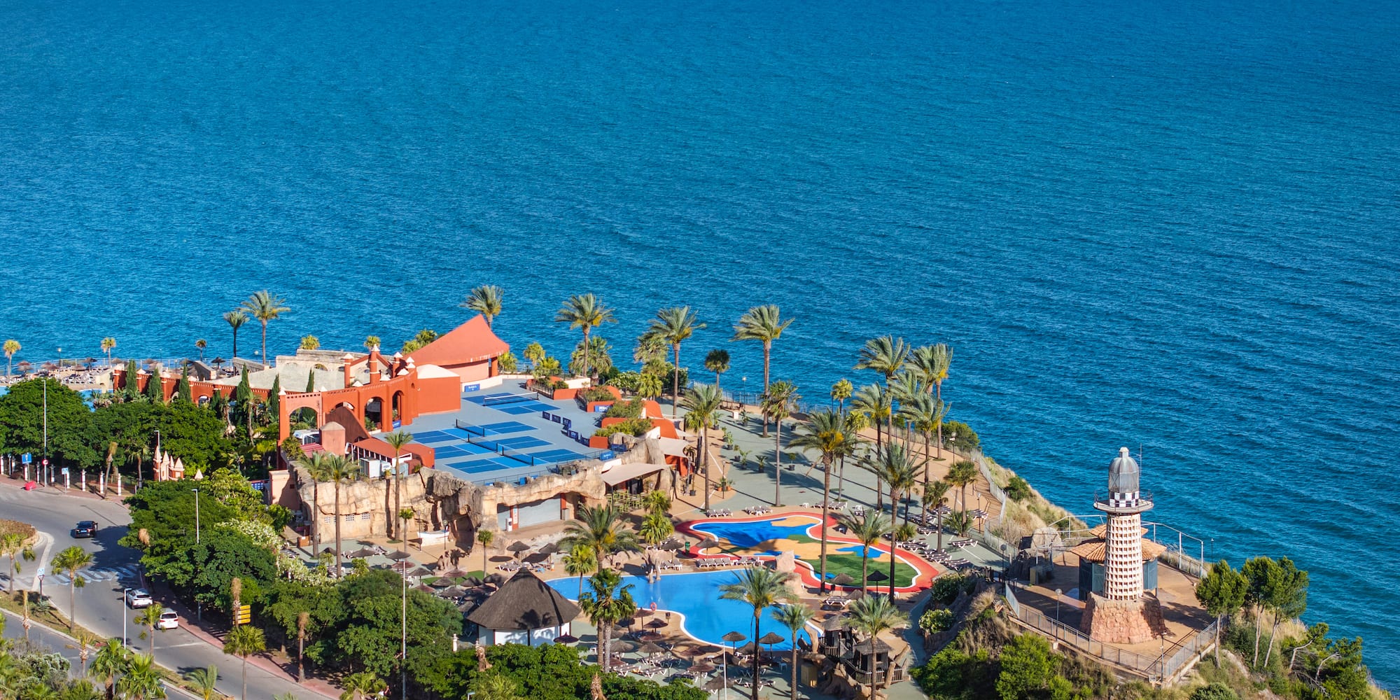 Coastal resort with palm trees, swimming pools, lighthouse, and blue sea.