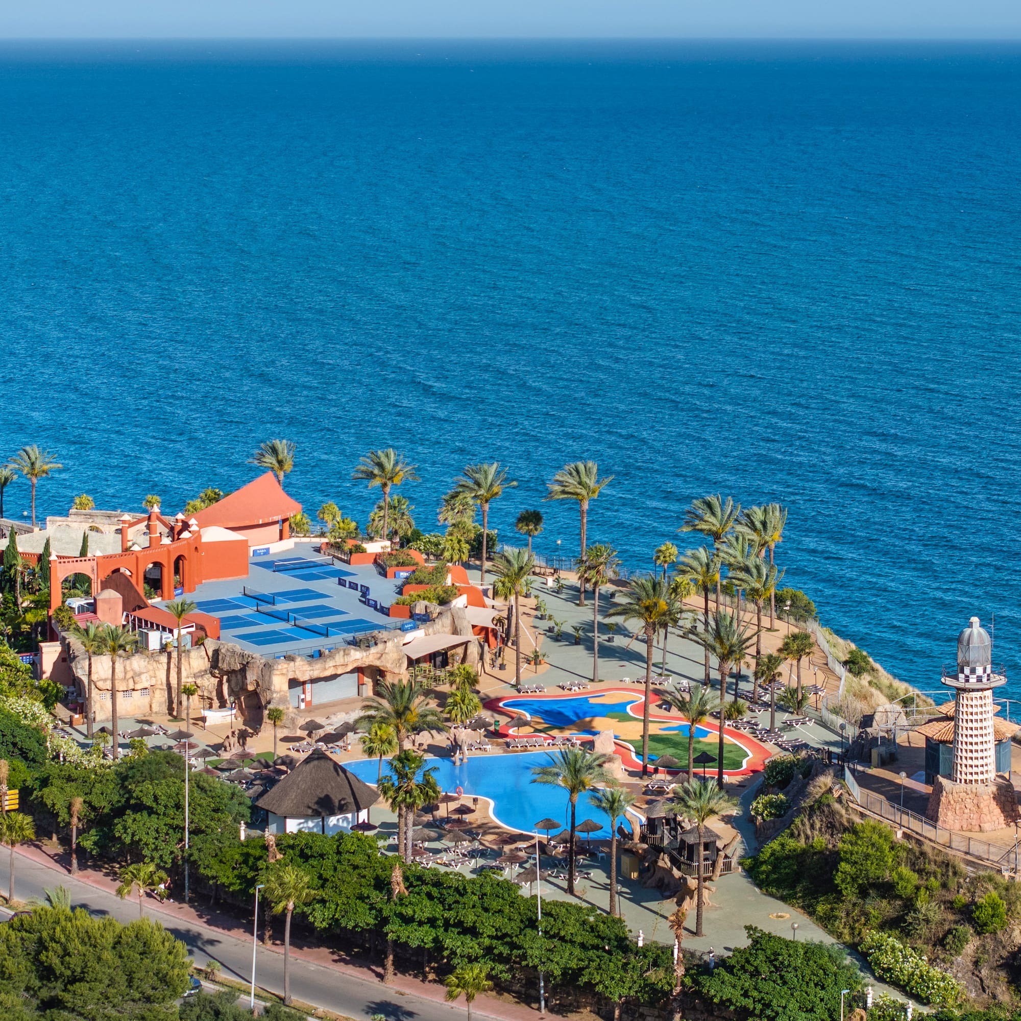Coastal resort with pools, palm trees, and lighthouse overlooking blue ocean.