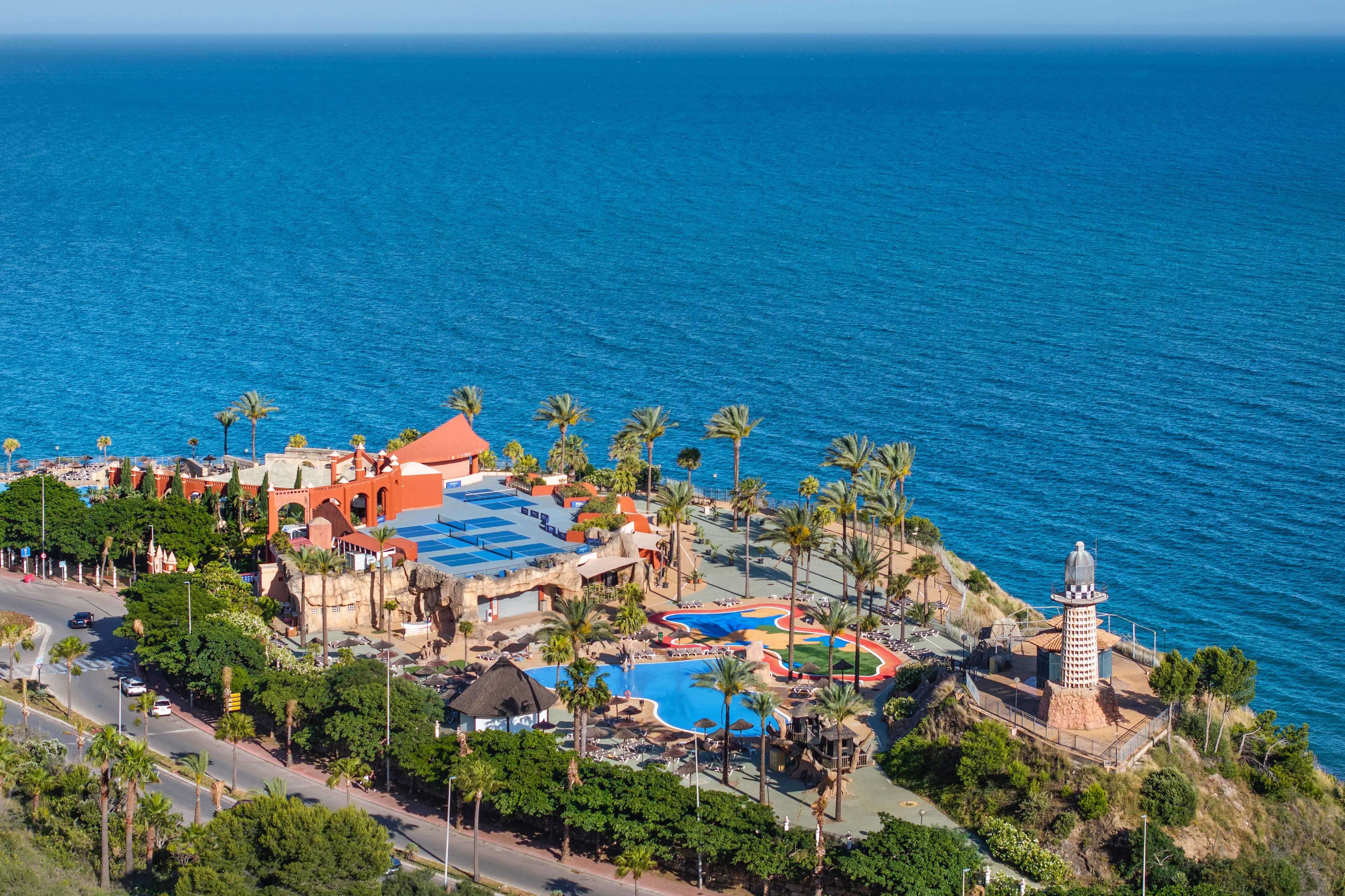 Coastal resort with pools, palm trees, and a lighthouse by the blue ocean.