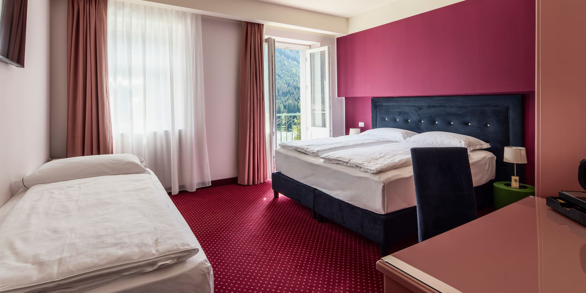 Comfortable hotel room with queen and single beds, red carpet, balcony mountain view.