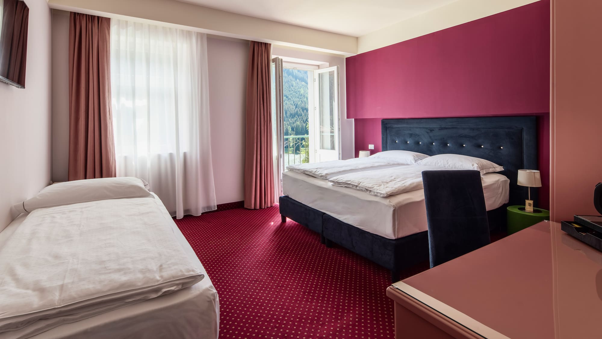 Hotel room with double and single beds, red carpet, and mountain view balcony.