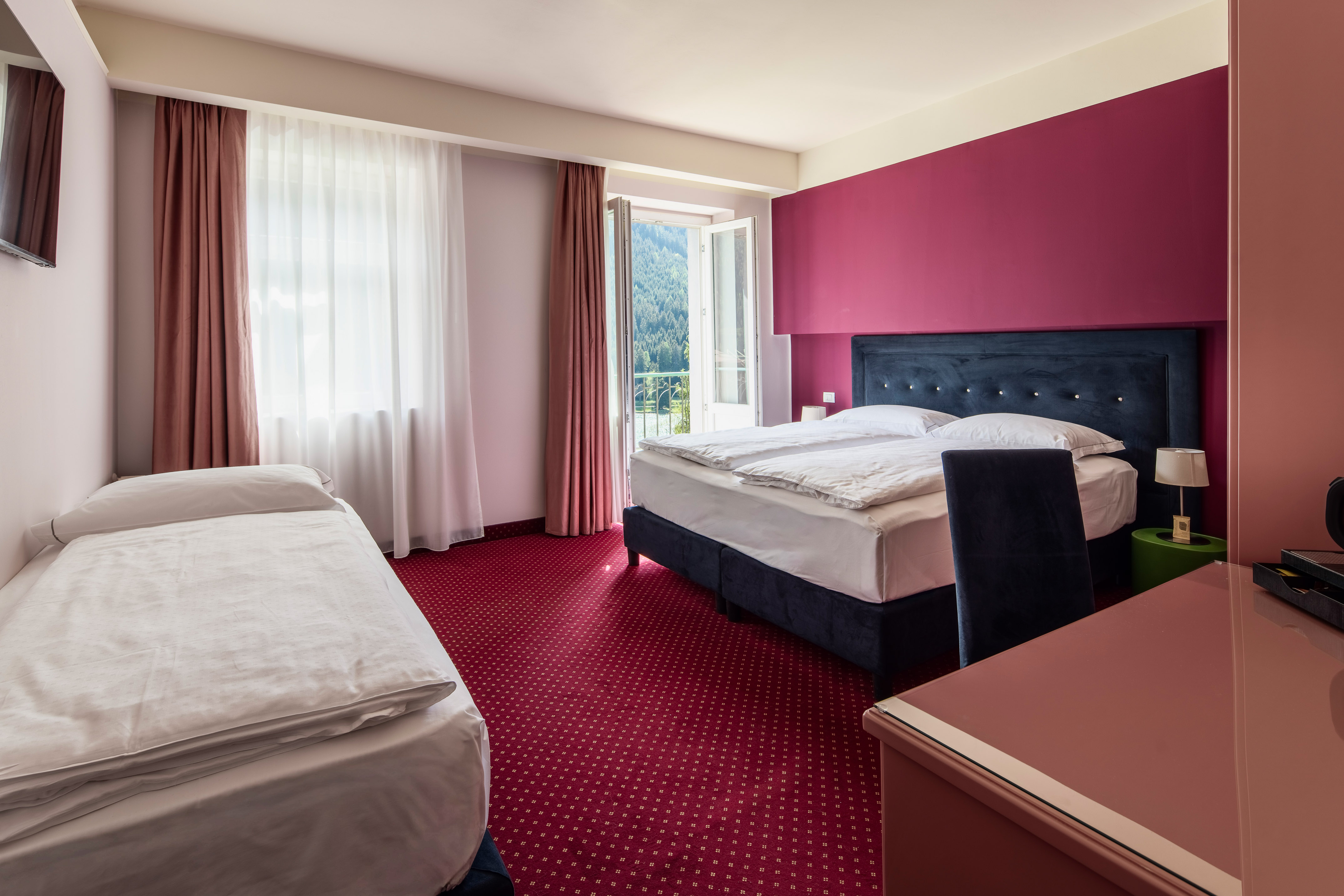 Comfortable hotel room with queen and single beds, red carpet, balcony mountain view.