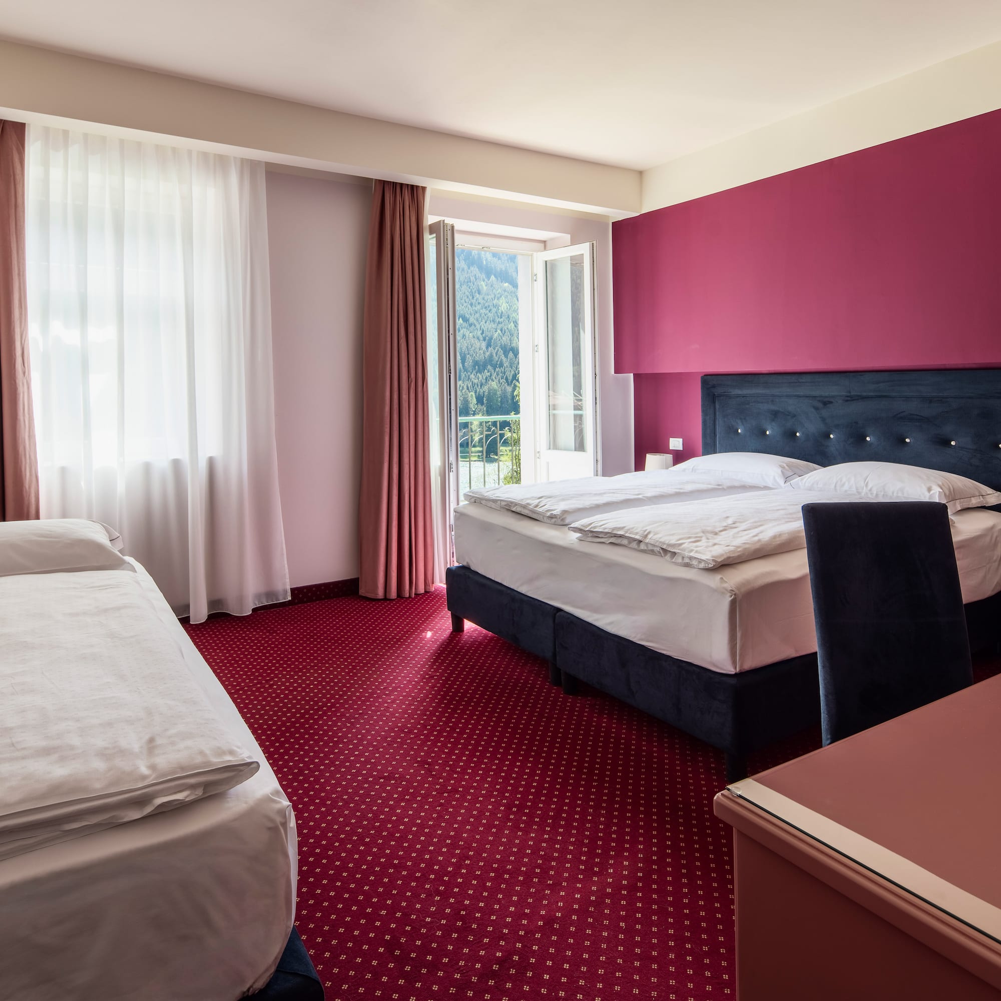 Comfortable hotel room with queen and single beds, red carpet, balcony mountain view.