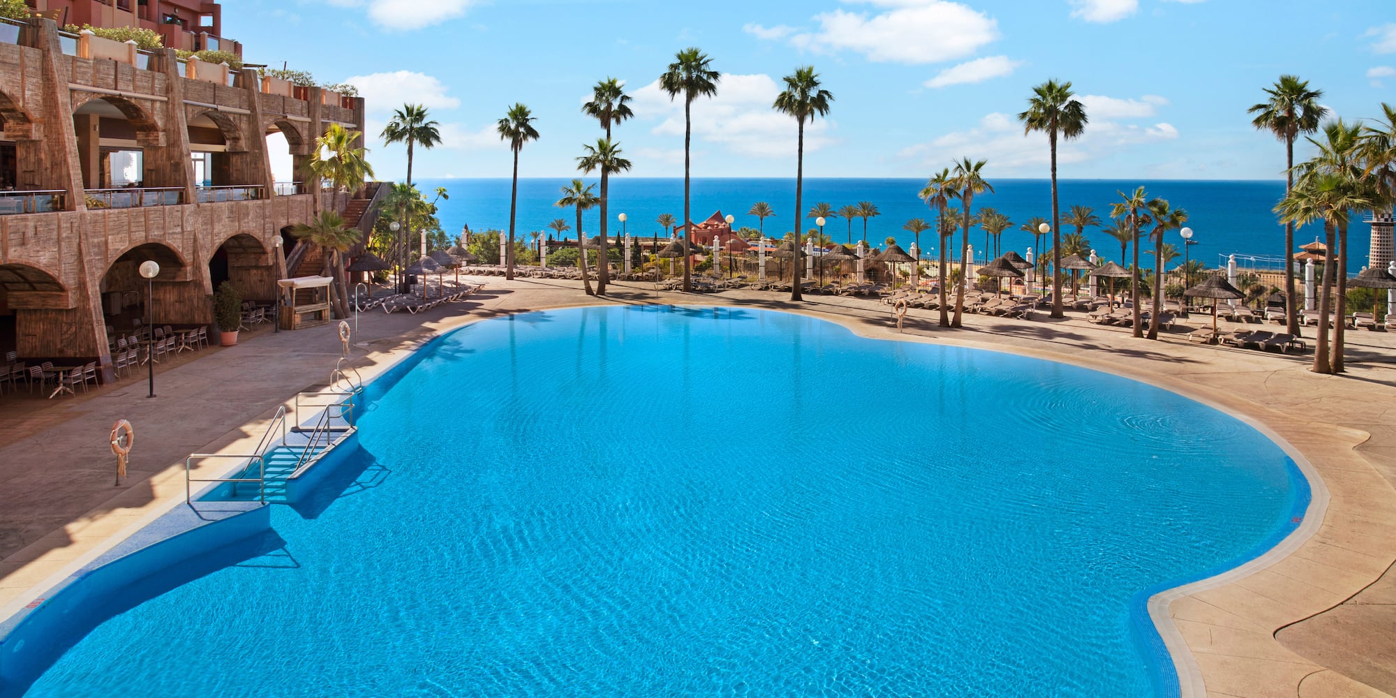 Large resort swimming pool with palm trees, ocean view, and hotel building.