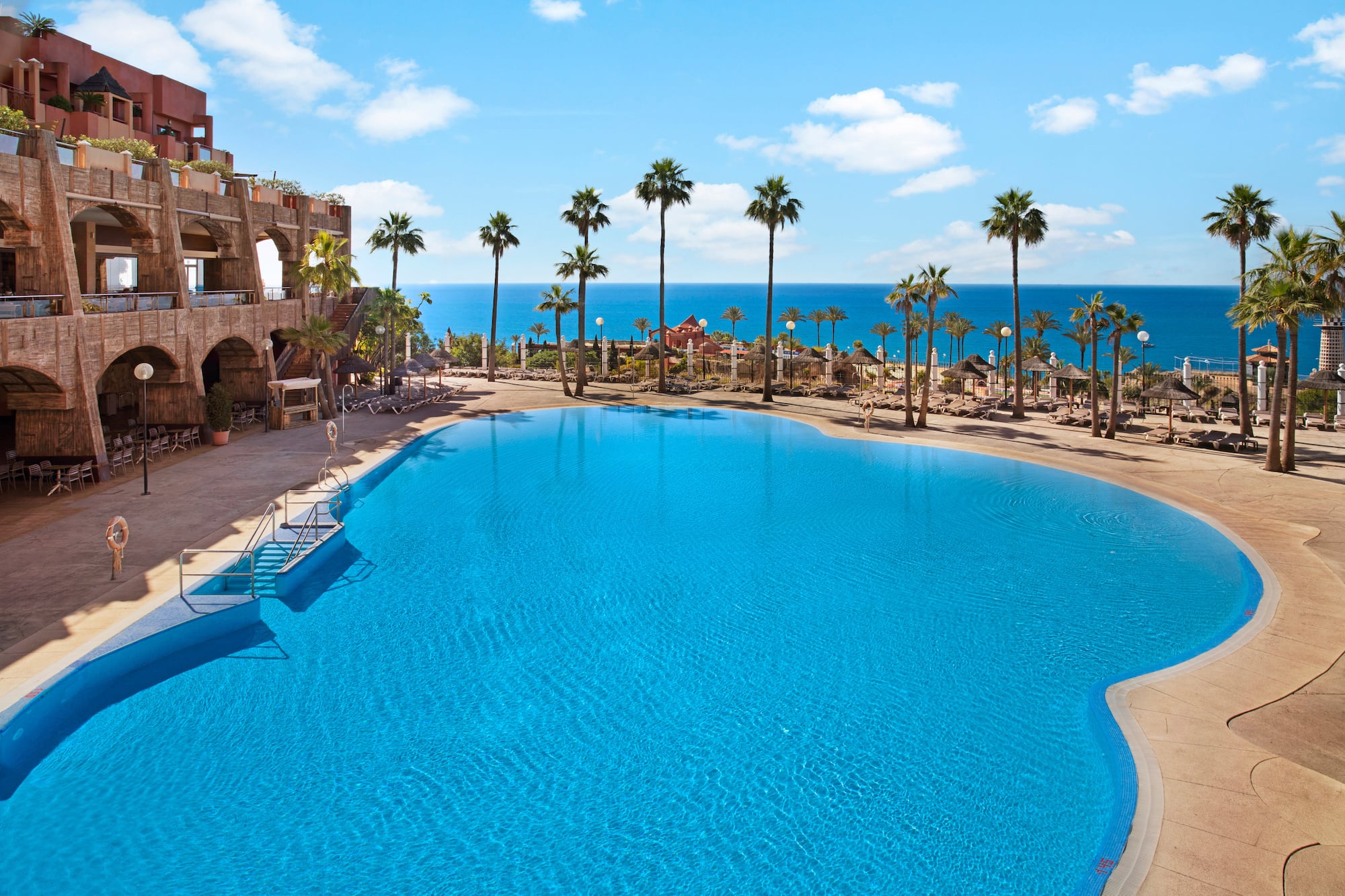 Resort swimming pool with ocean view, palm trees, and hotel.