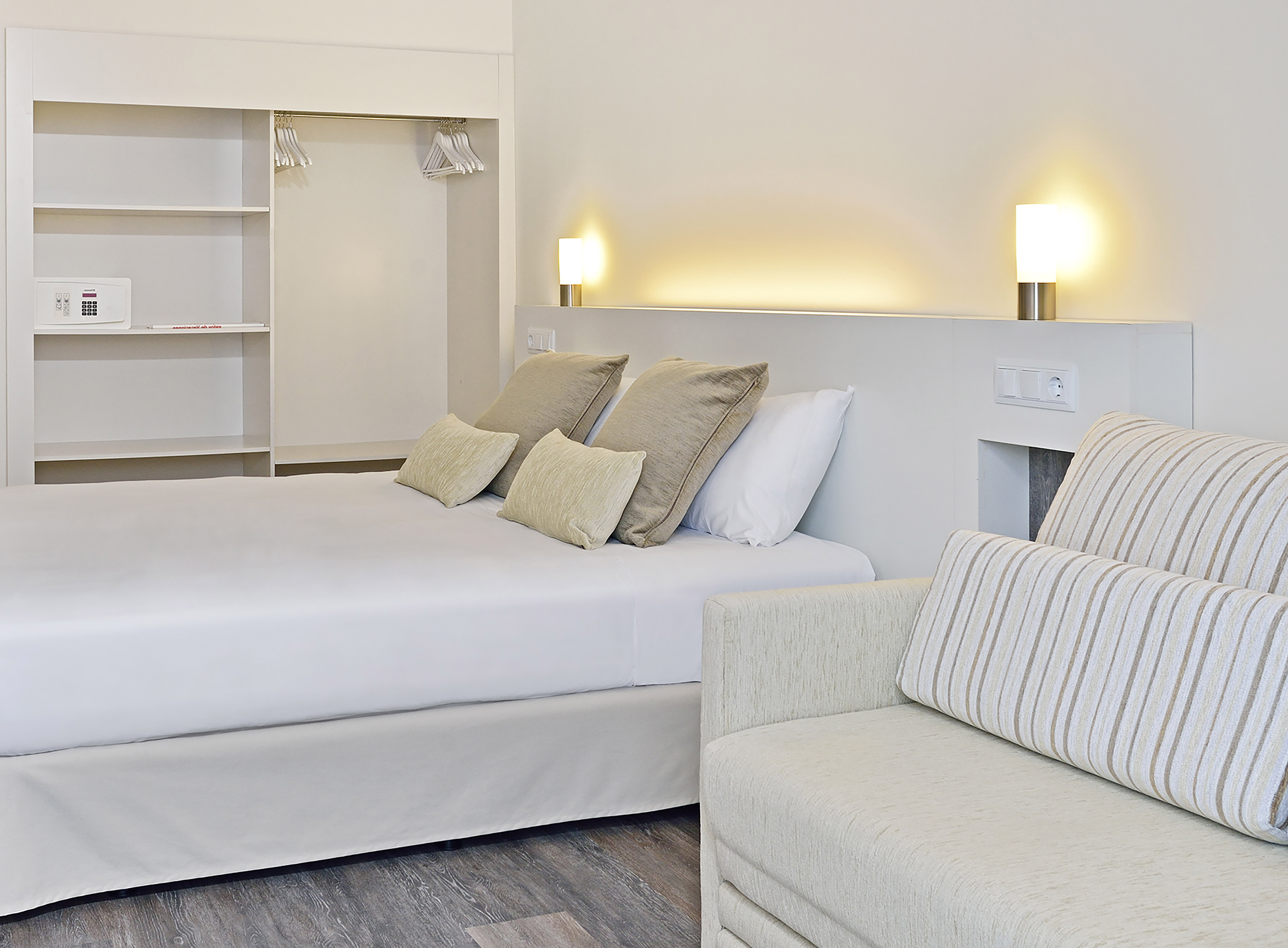 Modern hotel bedroom with white bed, built-in wardrobe, safe, and sofa.