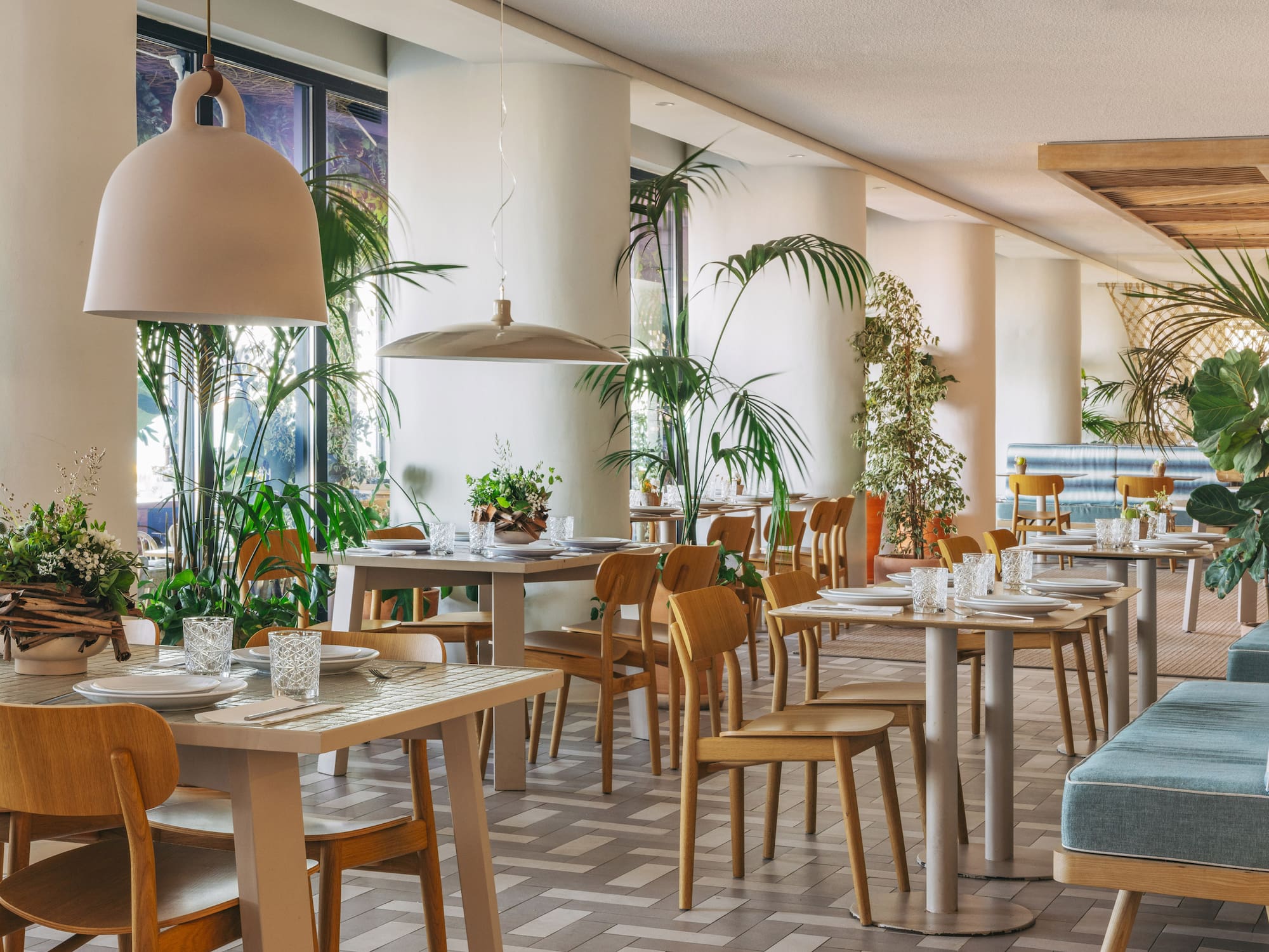 Modern restaurant interior with light wood tables, chairs, and lush tropical plants.