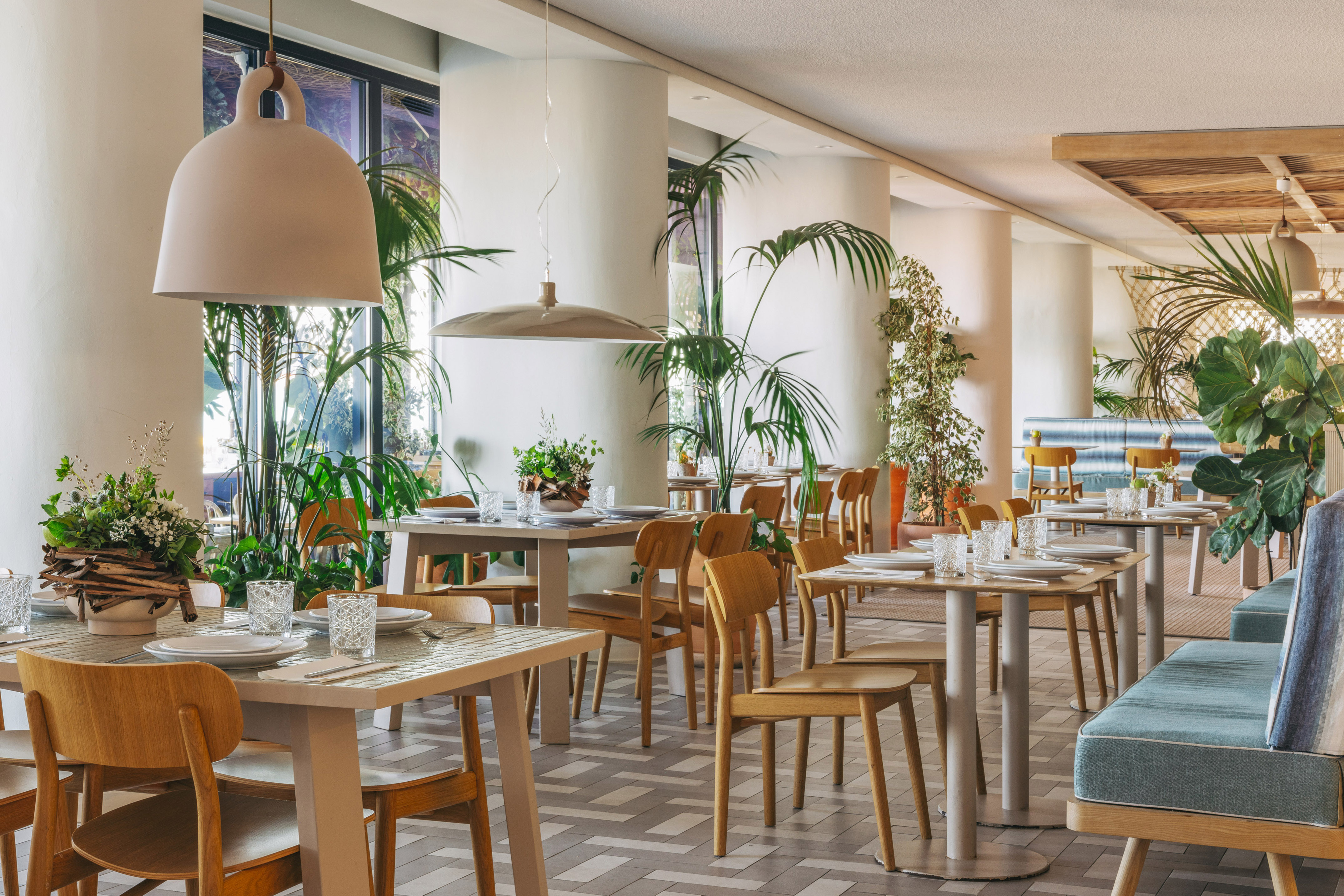Modern restaurant interior with light wood tables, chairs, and lush tropical plants.