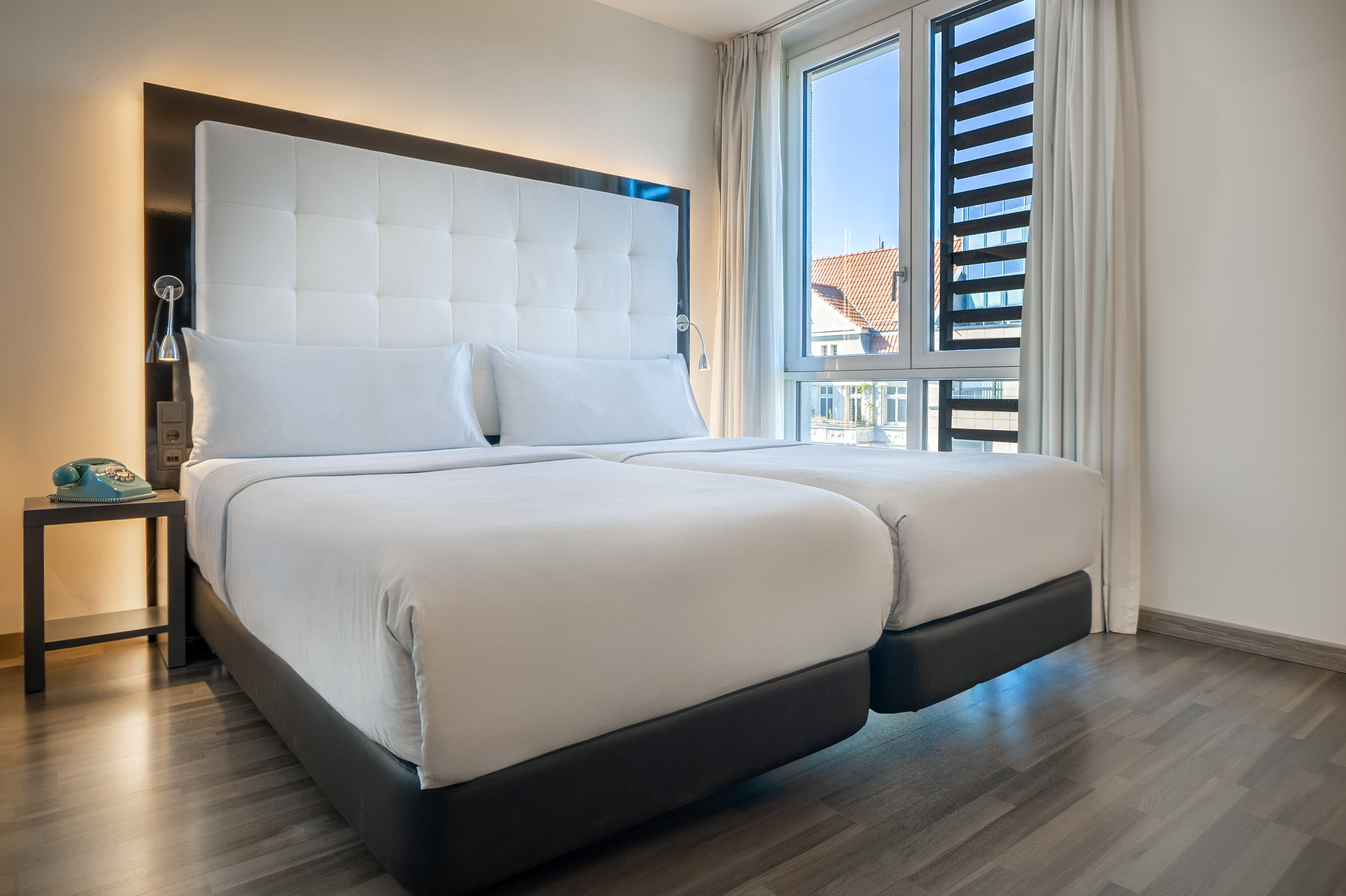 Modern hotel room with two white beds, tufted headboard, wood floor, and large window.