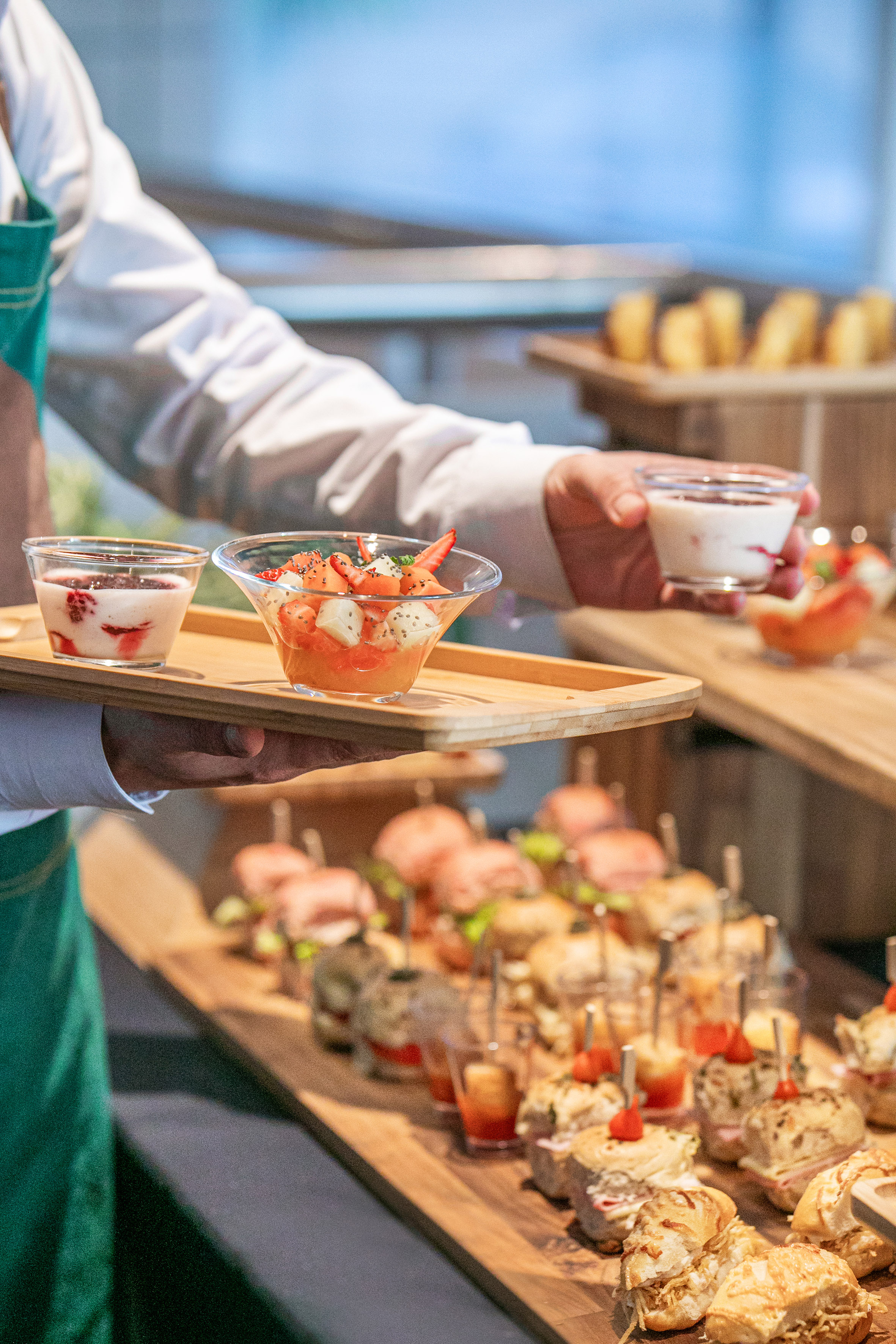 Catering staff holding fruit salad and yogurt desserts over a buffet table.