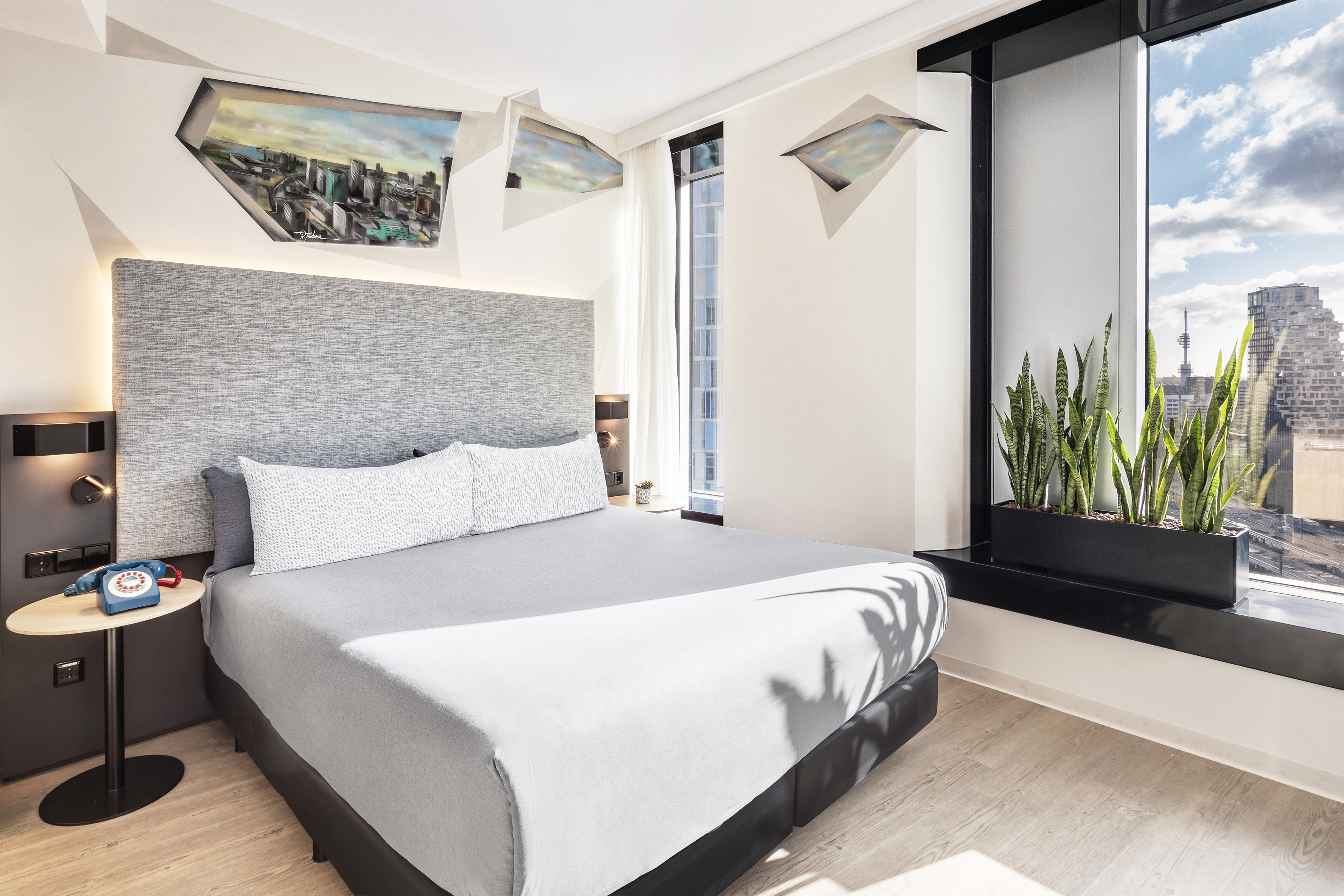 Modern hotel bedroom with king bed, city view window, snake plants, and abstract art.