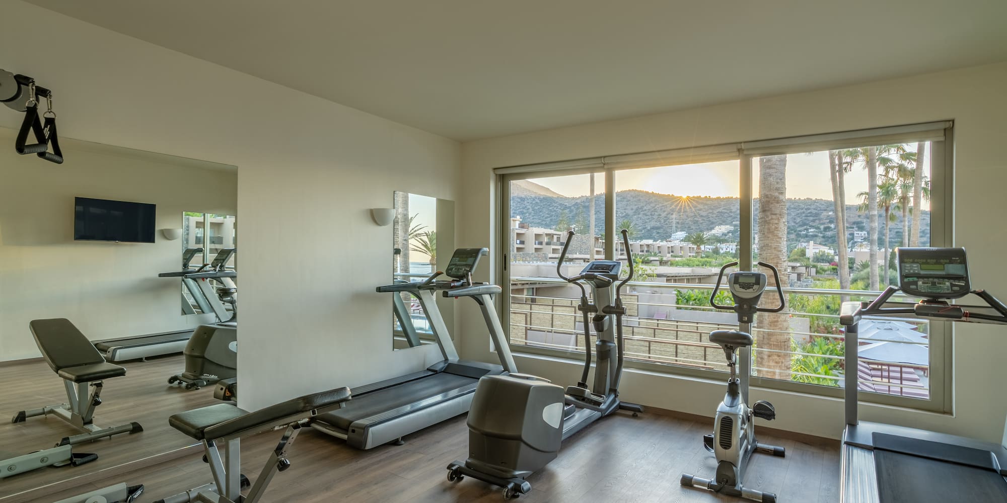Hotel gym with cardio equipment, weight bench, and sunset mountain view.