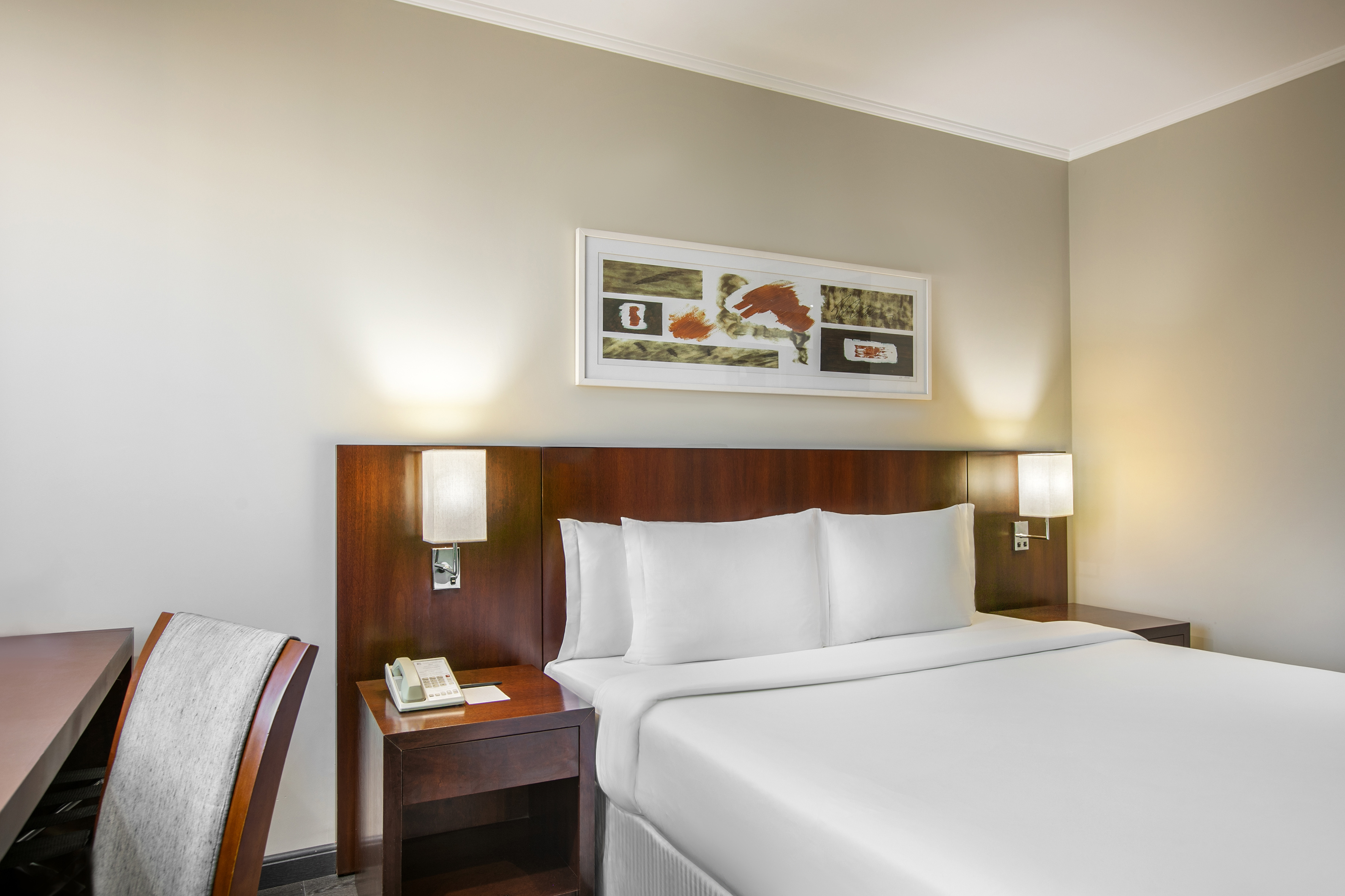 Modern hotel room with king bed, dark wood headboard, sconce lights, and abstract art.