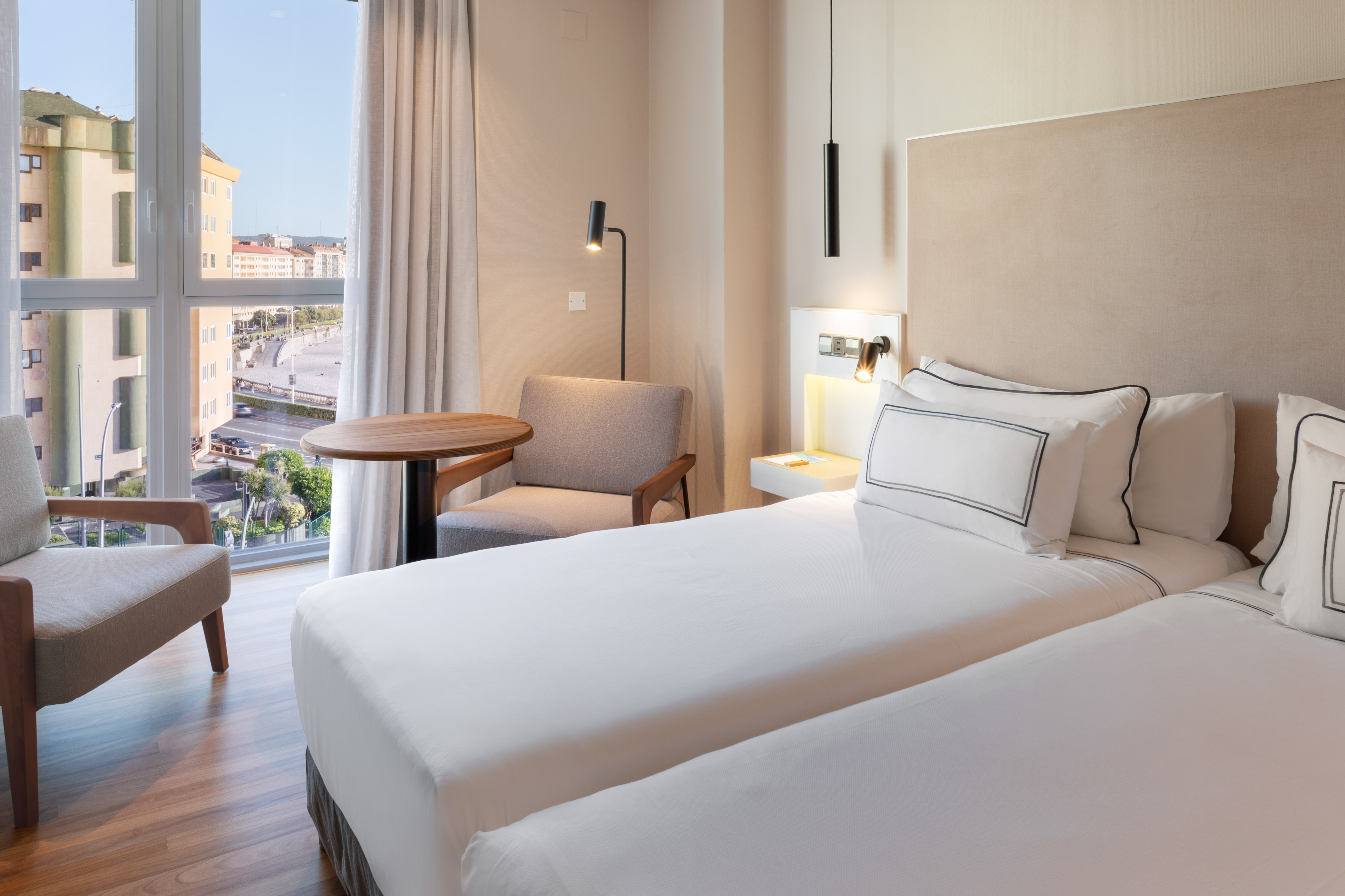 Modern hotel room, twin beds, city view window, armchair, wood floor.