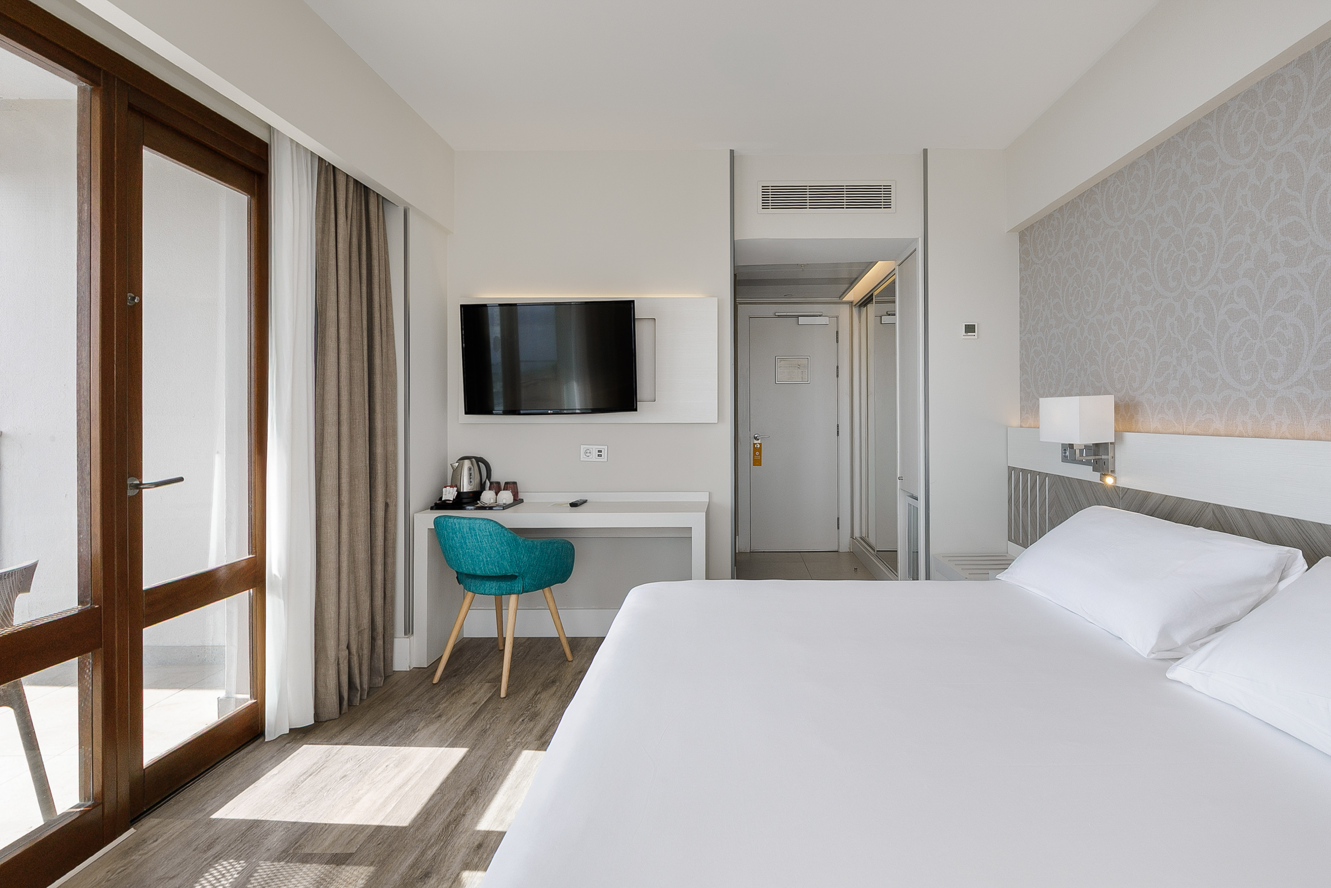 Bright hotel room with large bed, desk, TV, and balcony access.