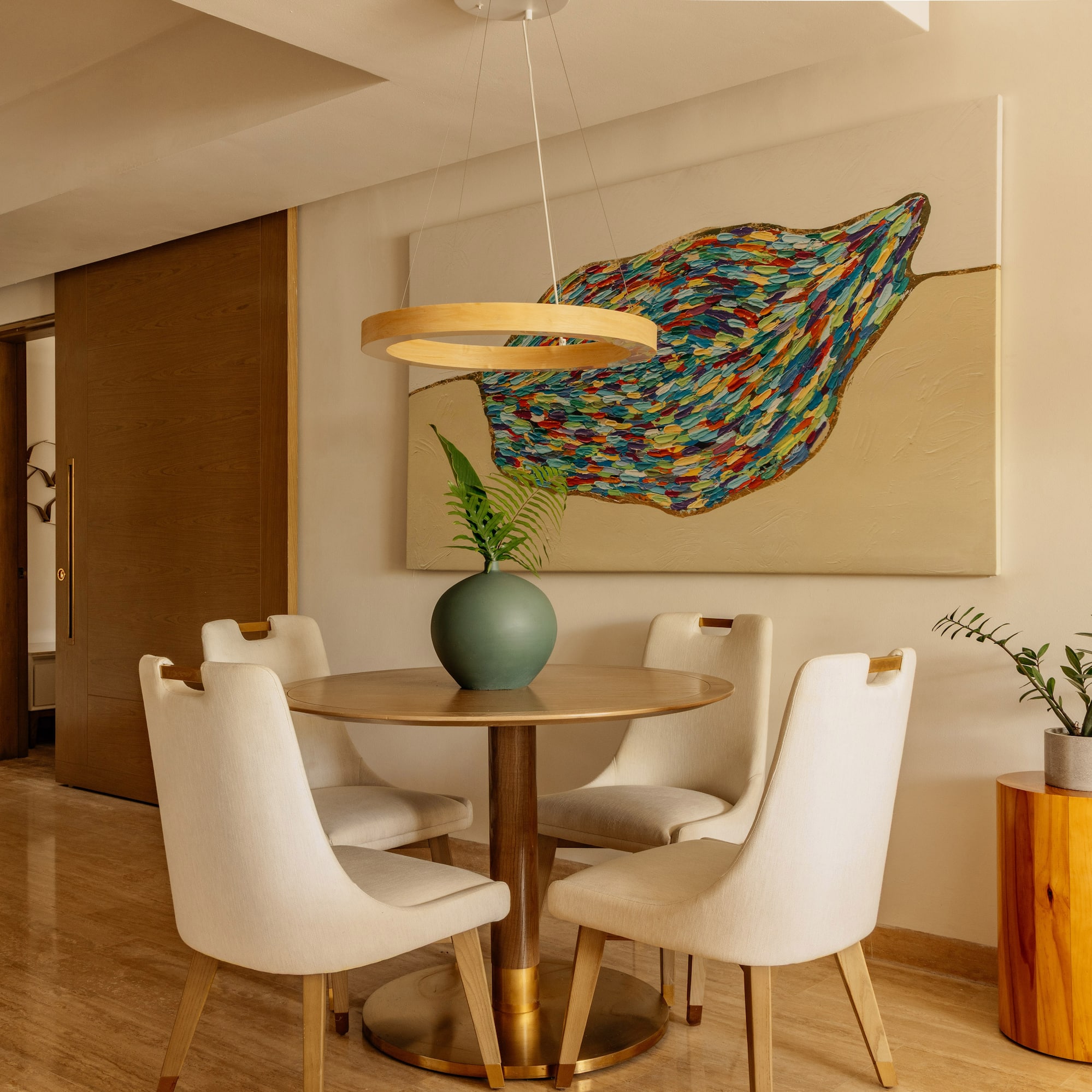 Modern dining room with round wood table, cream chairs, abstract art, and plants.