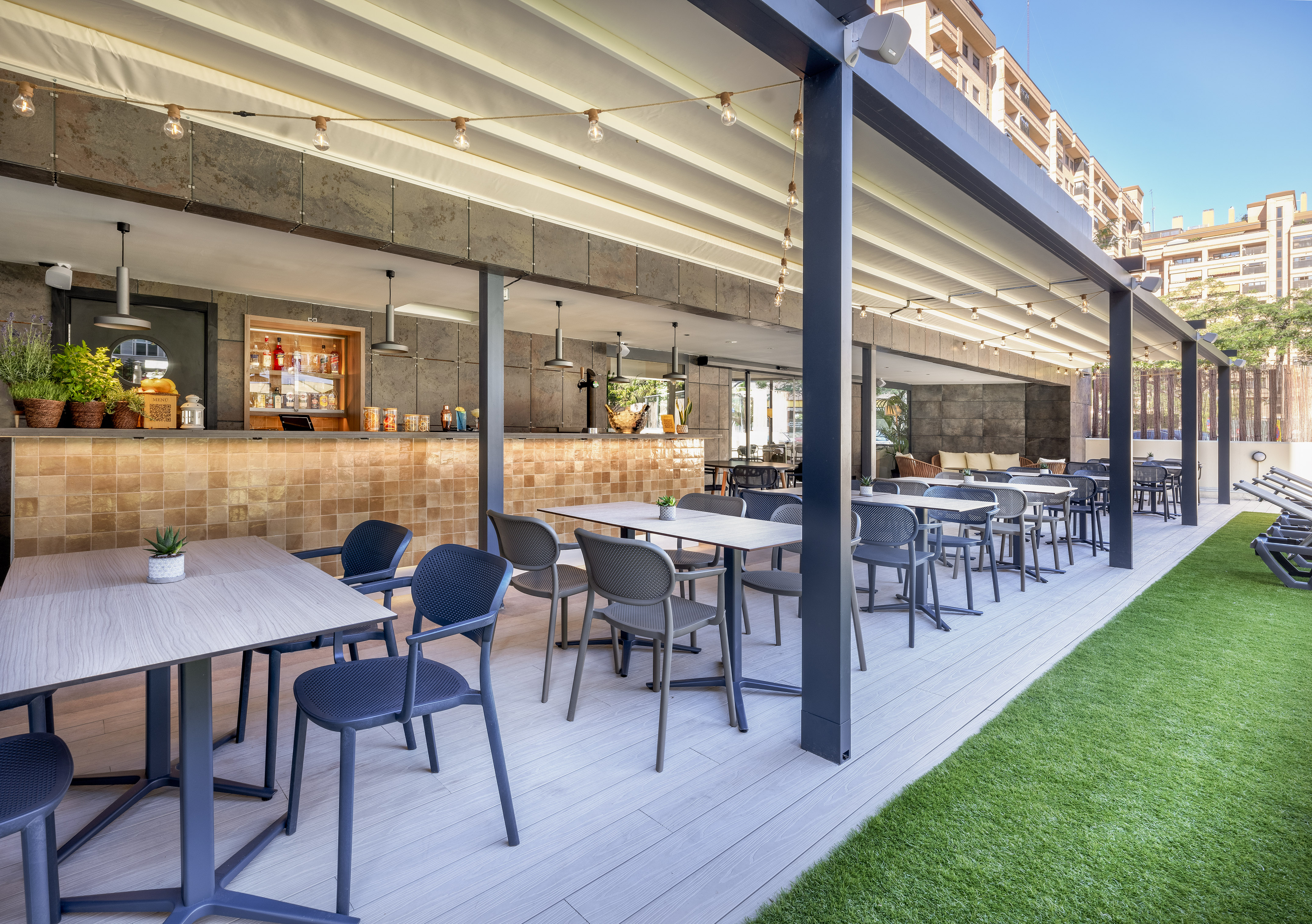 Modern outdoor bar and dining patio with tables, canopy, string lights, and turf.