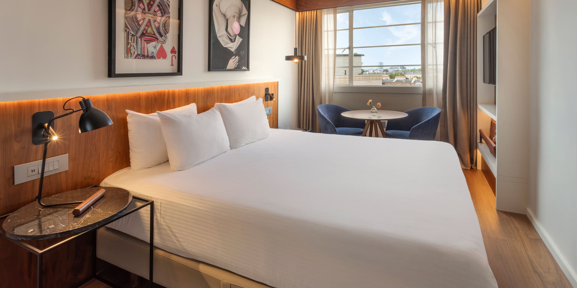 Modern hotel room with white bed, framed art, cityscape window, and seating.