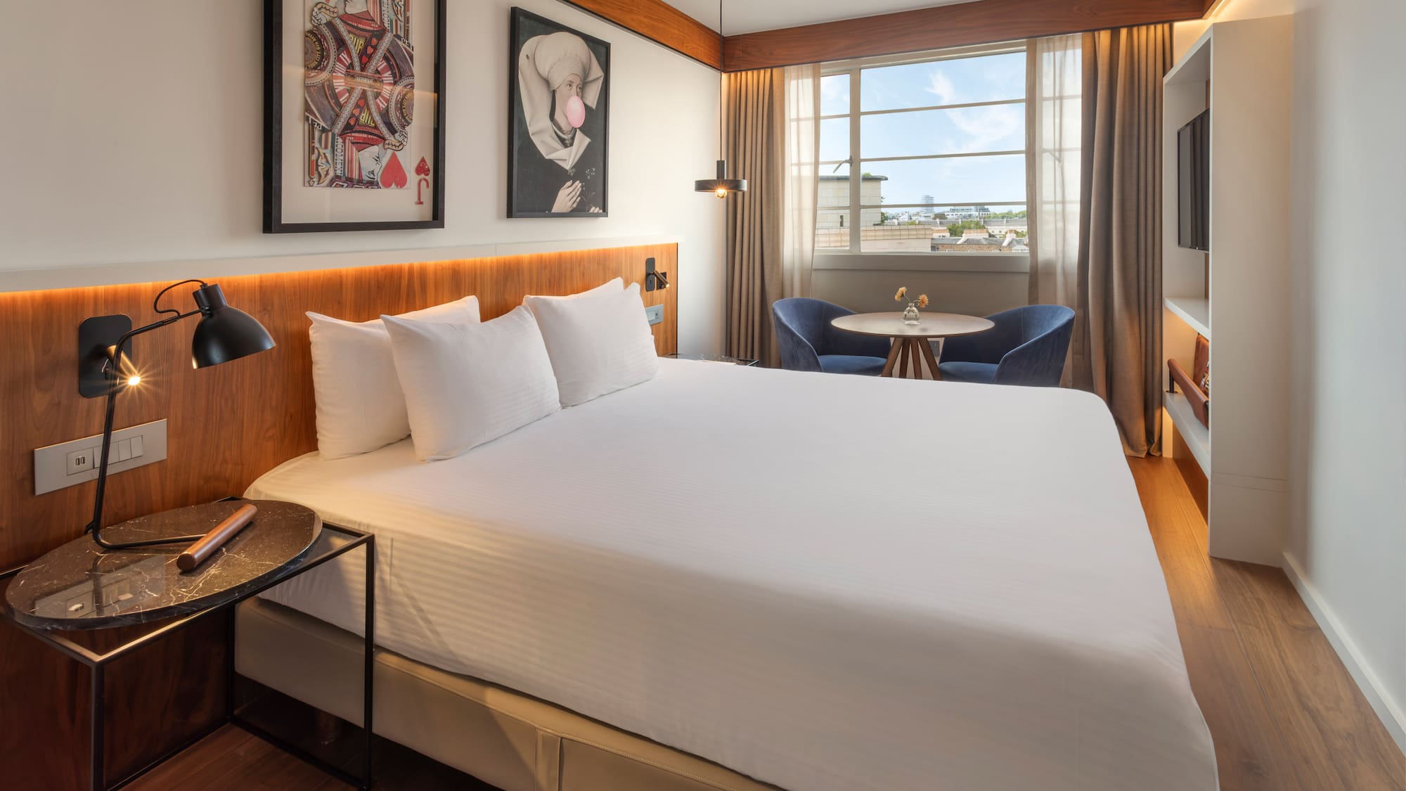 Modern hotel room with white bed, framed art, cityscape window, and seating.
