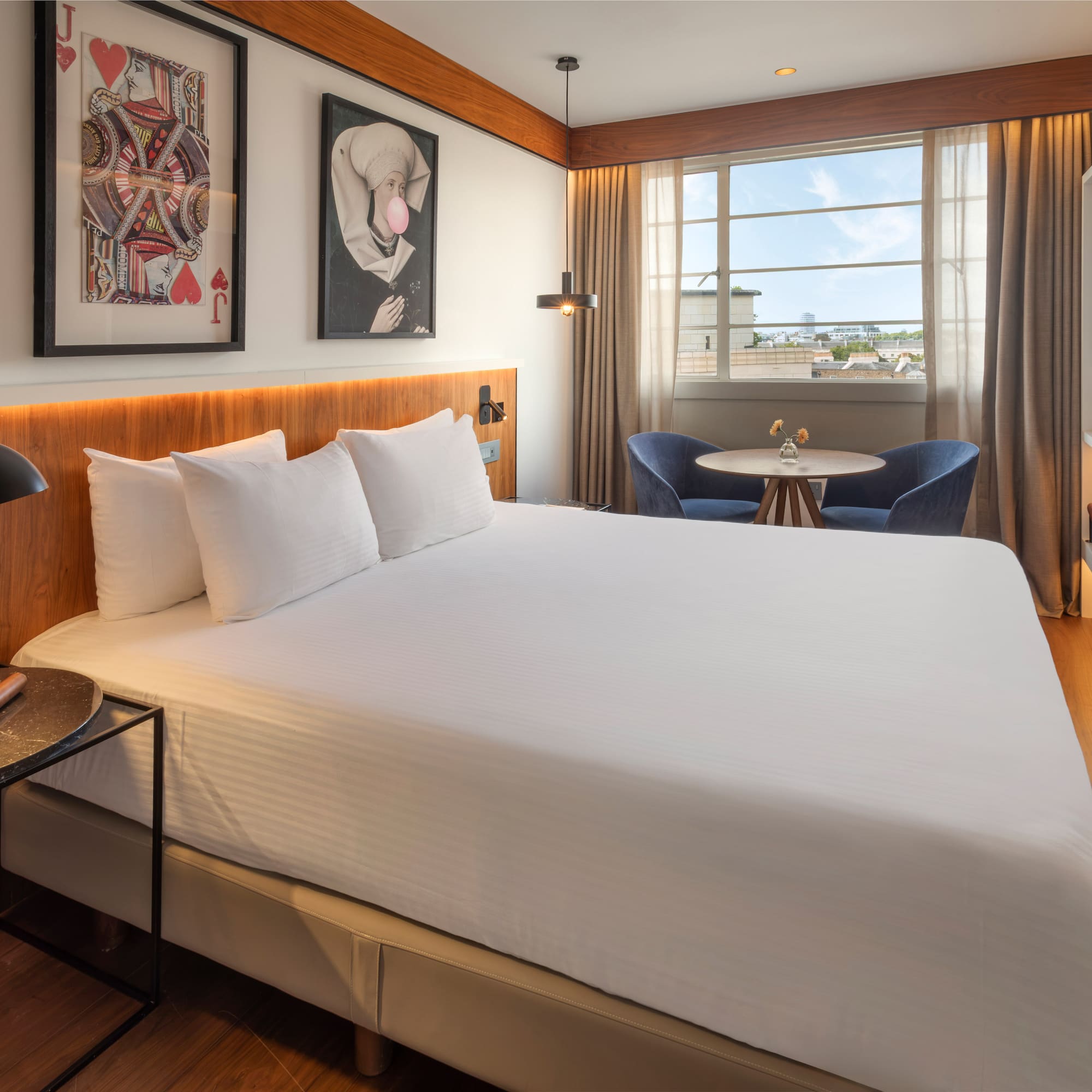 Modern hotel room with white bed, framed art, cityscape window, and seating.