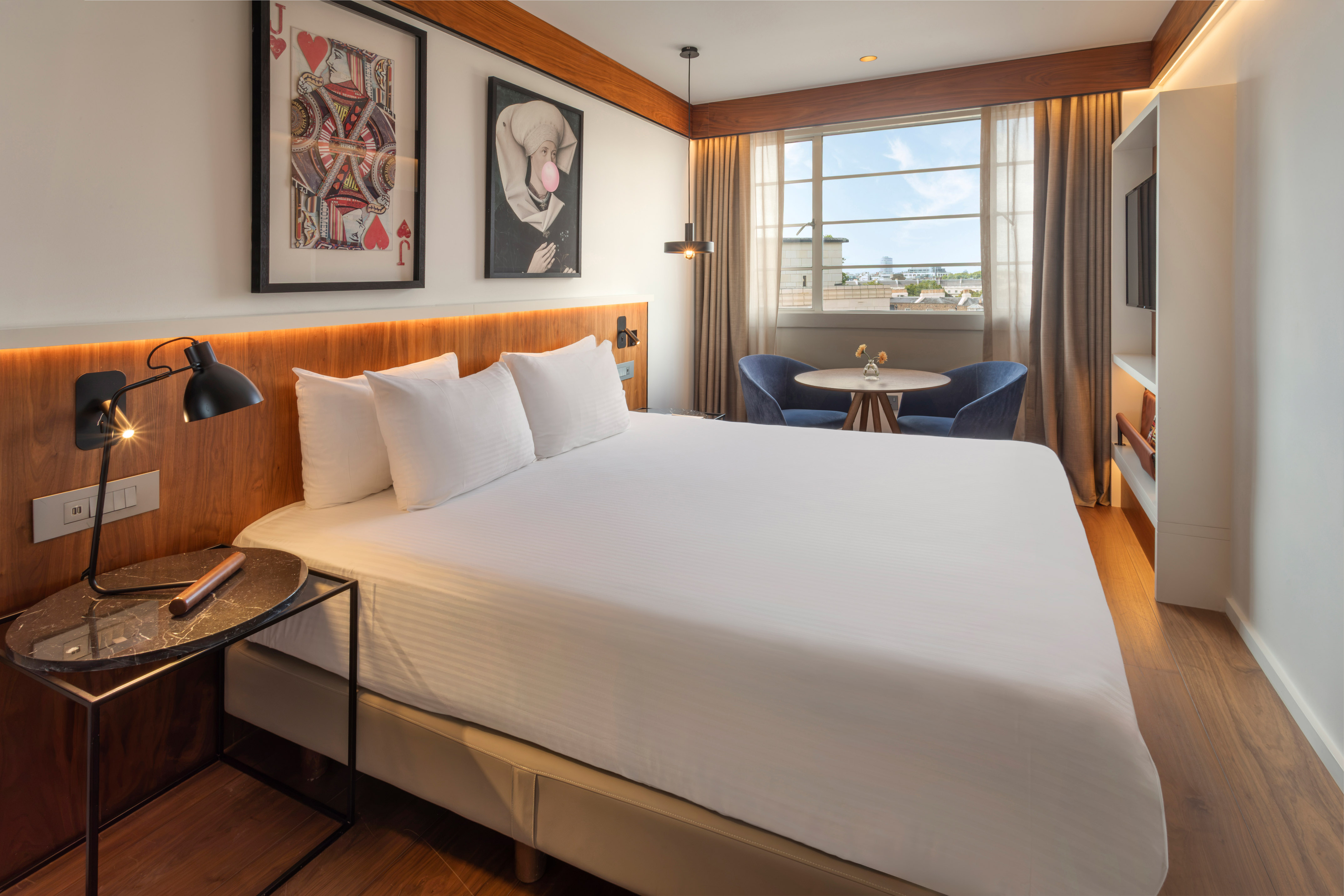Modern hotel room with white bed, framed art, cityscape window, and seating.