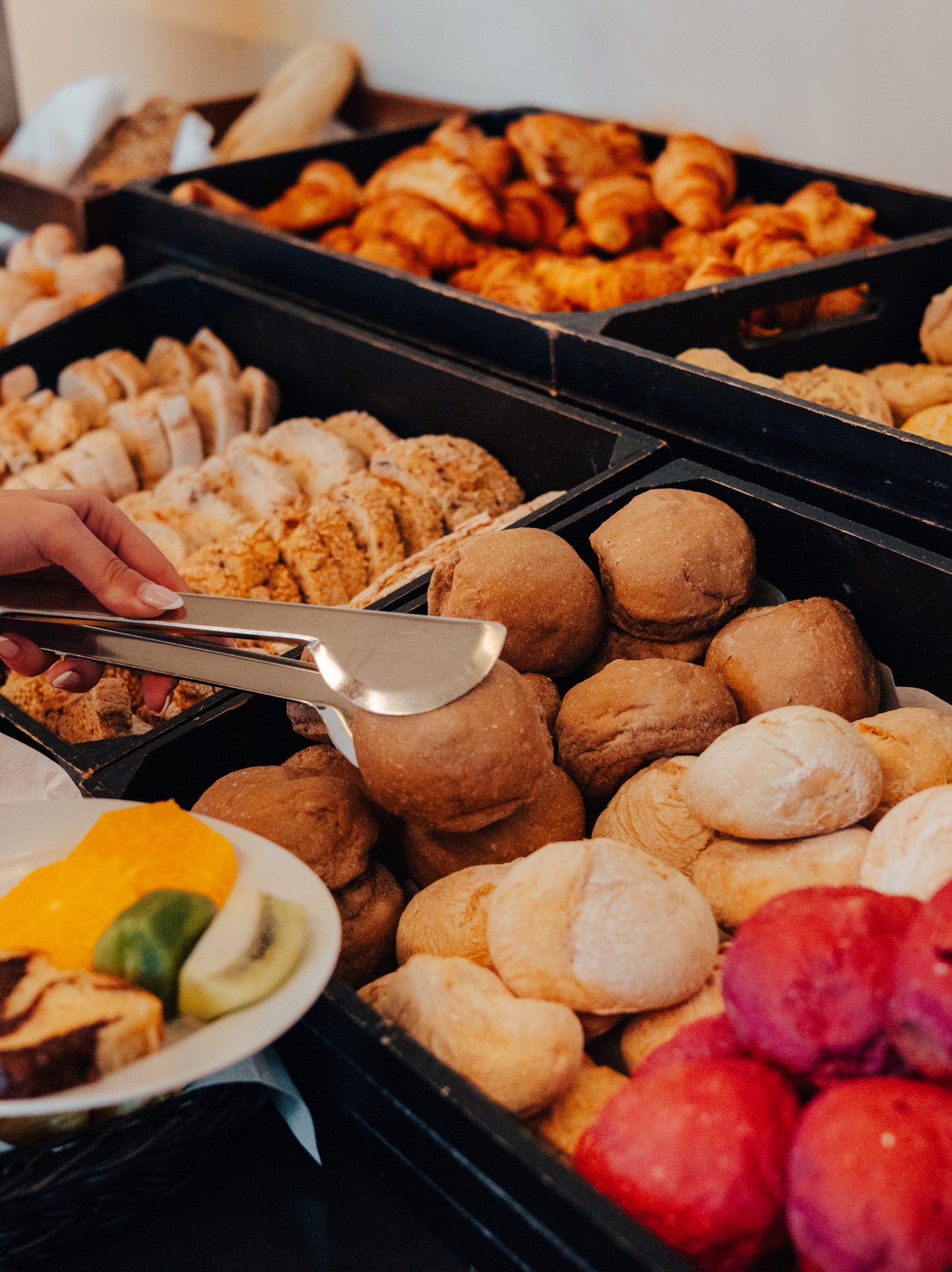 Hands serving diverse bread, croissants, and fruit from a breakfast buffet.