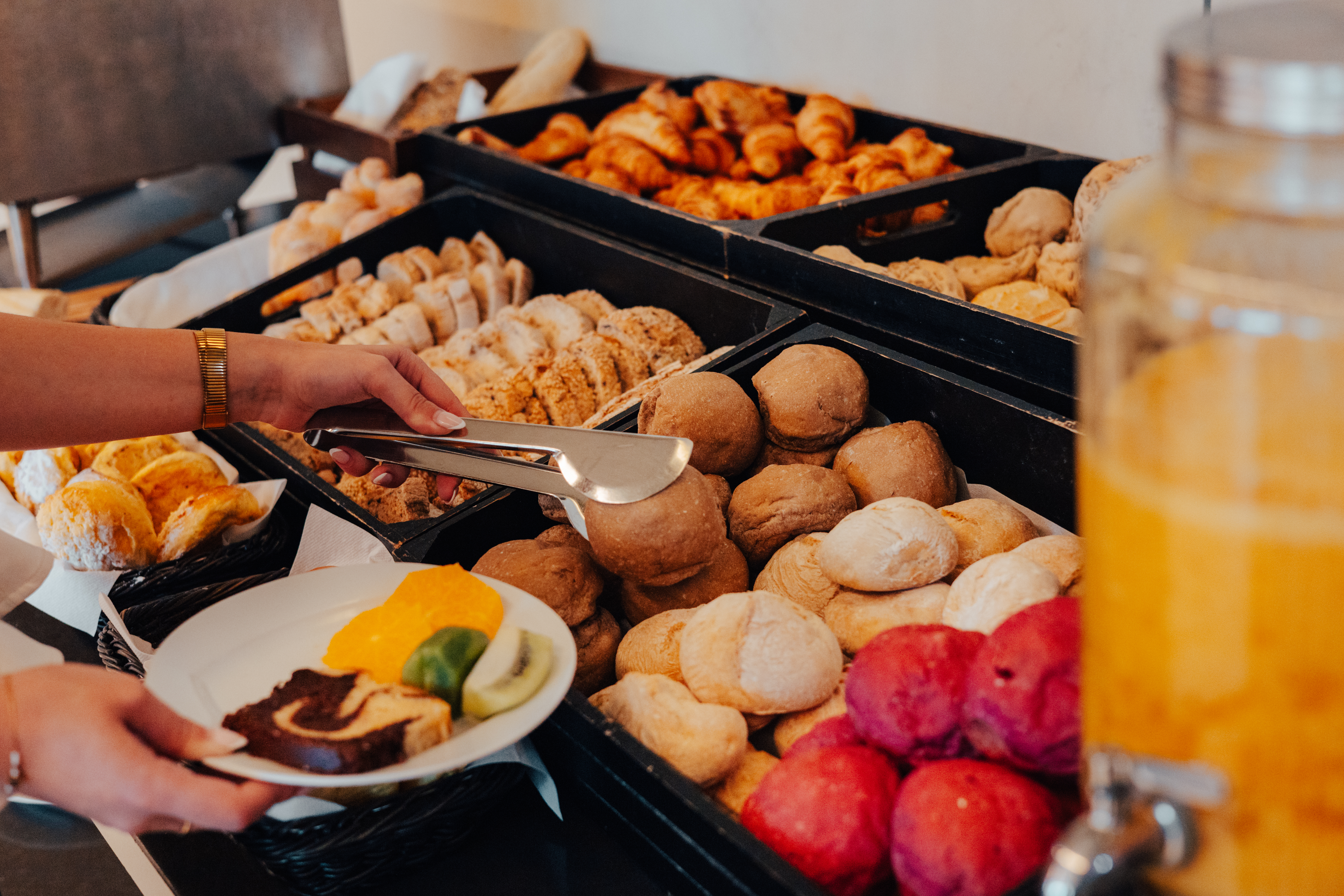 Hands serving diverse bread, croissants, and fruit from a breakfast buffet.