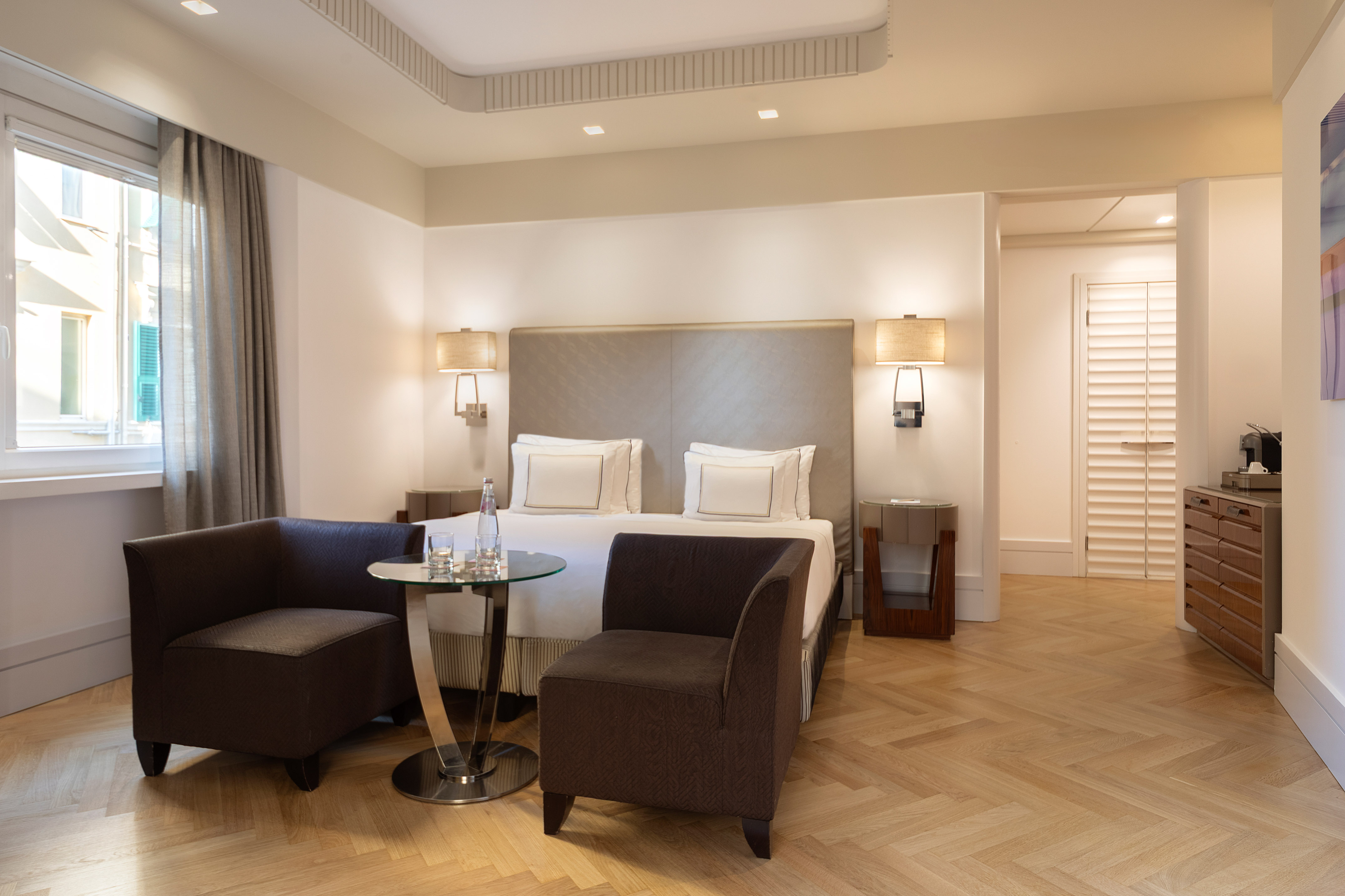 Elegant hotel bedroom with king bed, brown armchairs, and herringbone parquet floor.