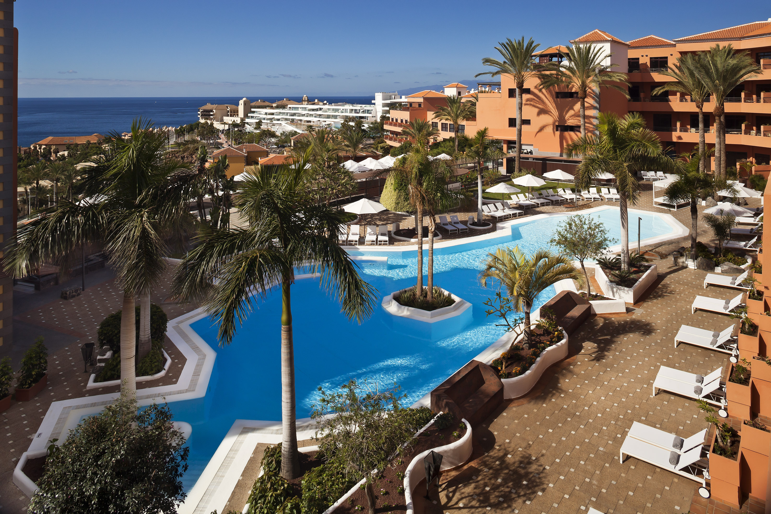 Tropical hotel pool with palm trees, orange resort buildings, and ocean view.