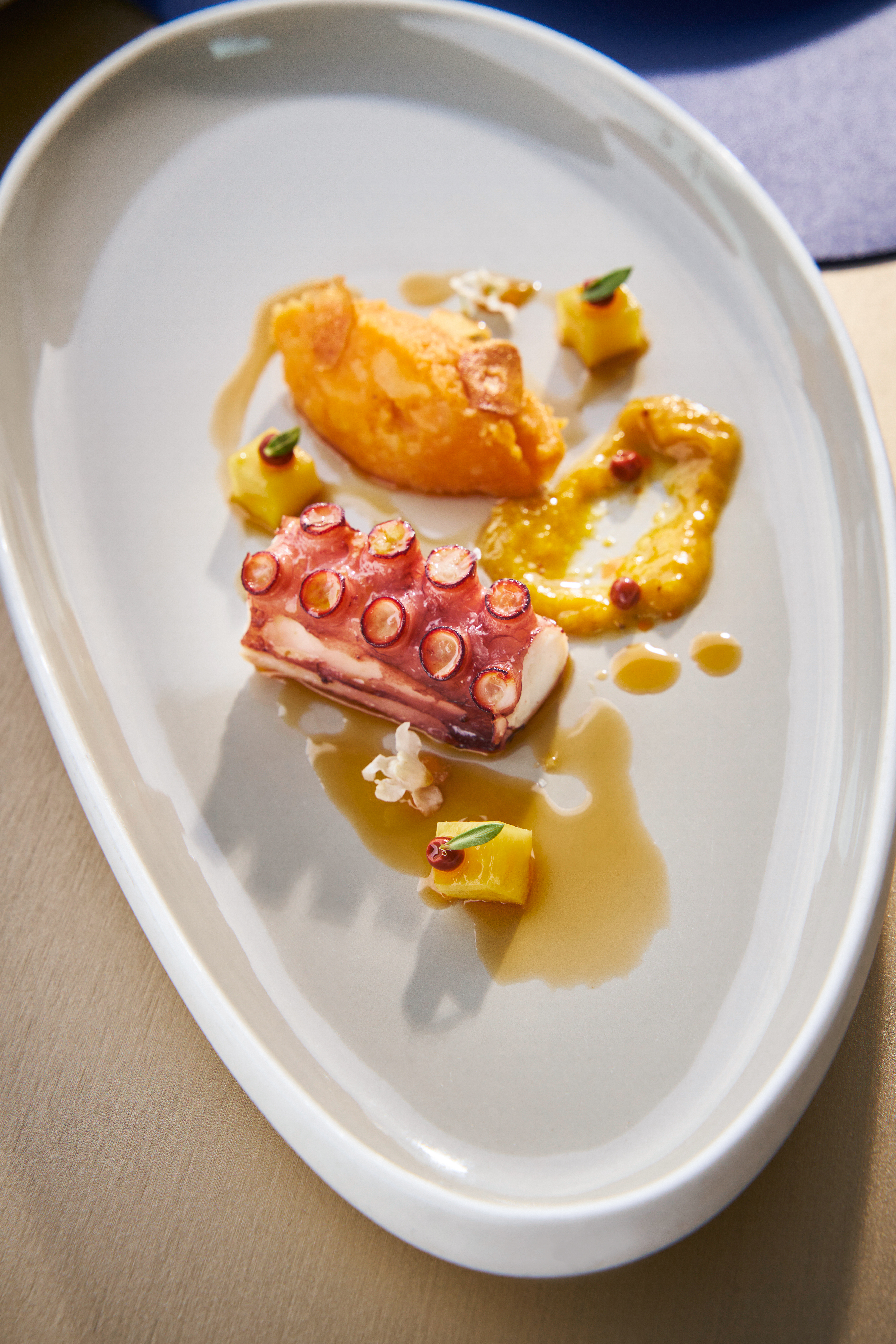 Cooked octopus tentacle, orange puree, pineapple, and sauce on a grey plate.