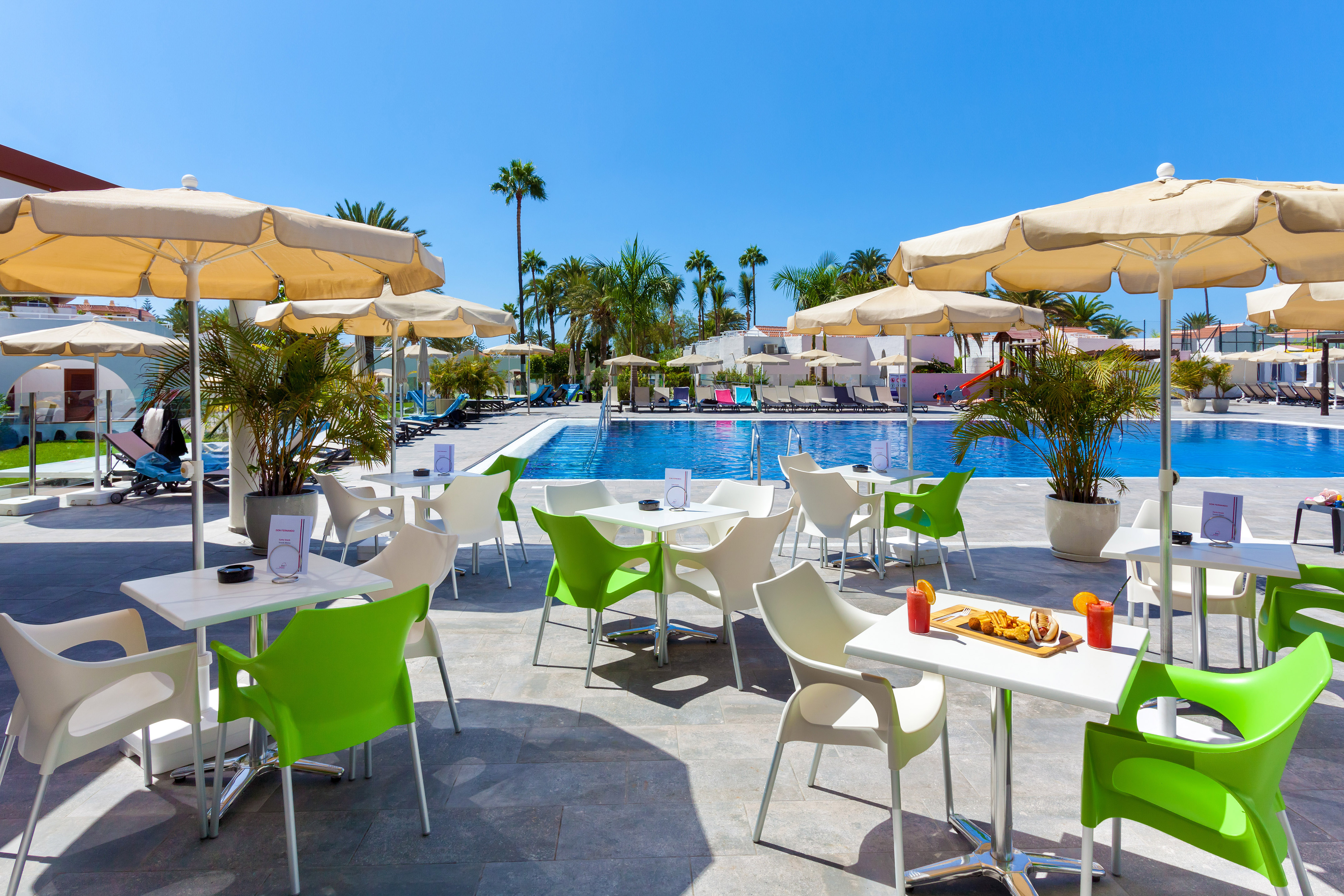 Sunny resort poolside cafe patio with green chairs and palm trees.