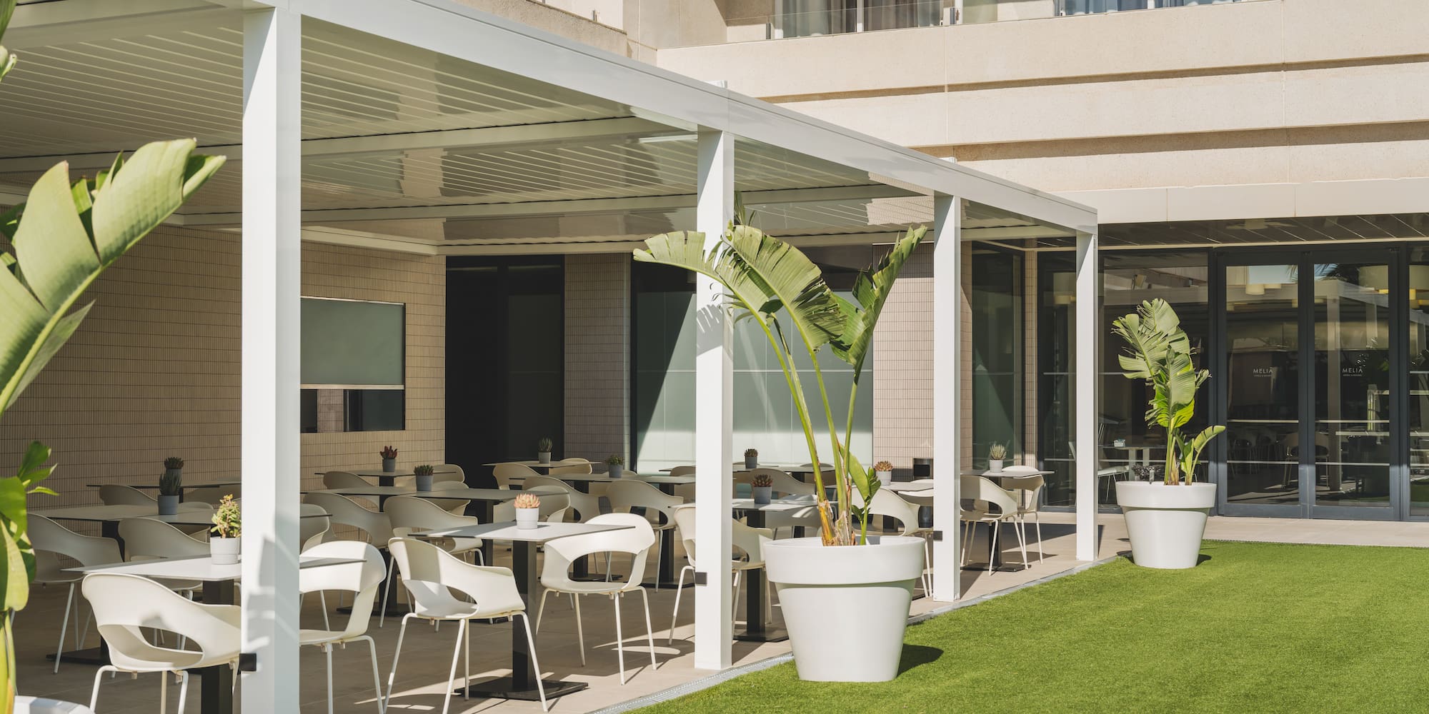 Terraza de hotel con pérgola blanca, mesas de exterior y césped artificial.