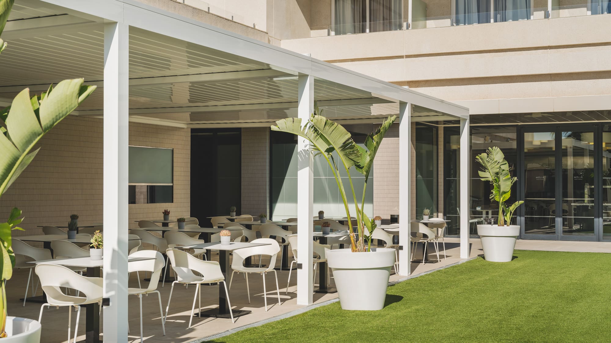 Terraza de hotel con pérgola blanca, mesas de exterior y césped artificial.