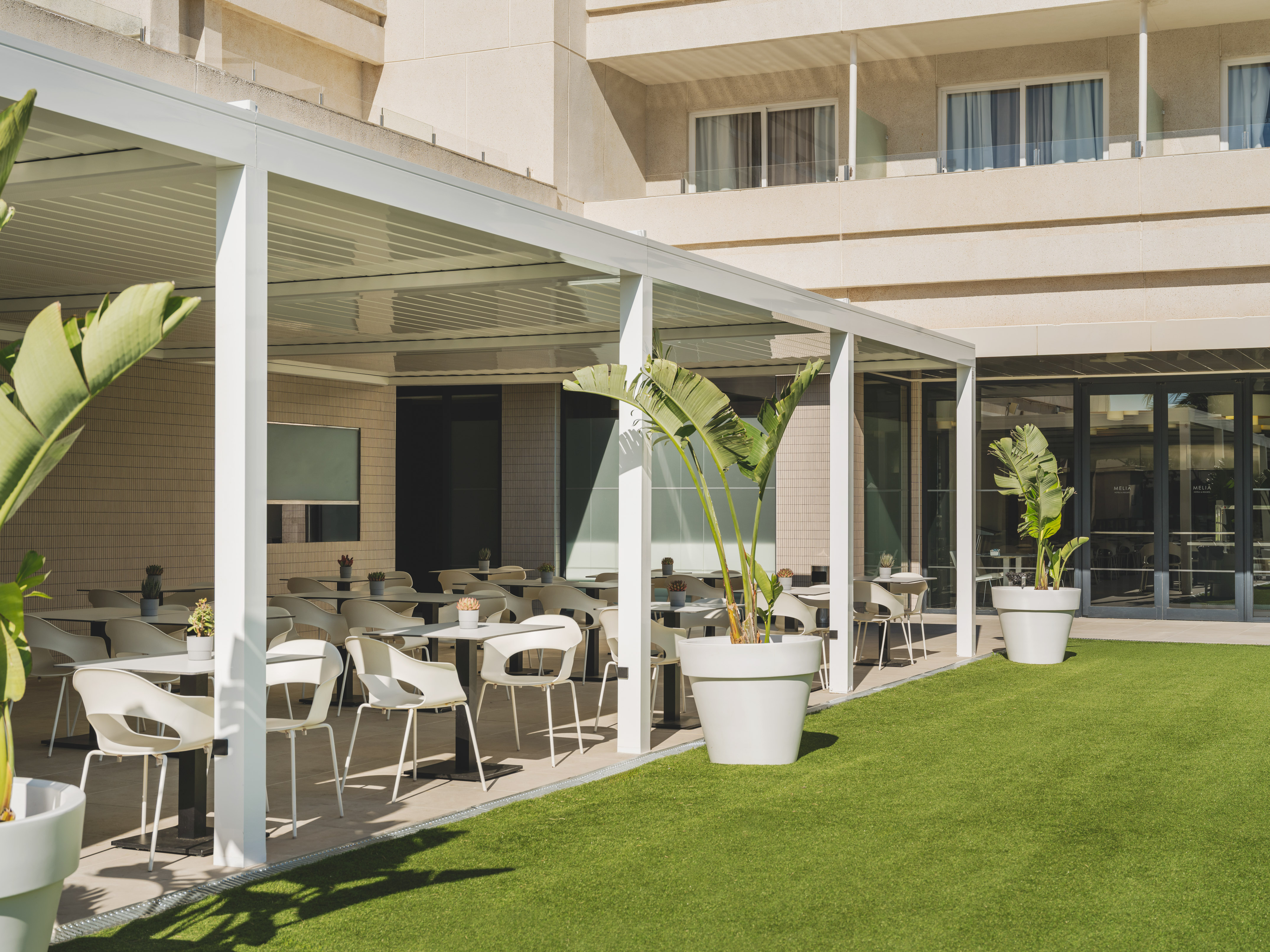 Terraza de hotel con pérgola blanca, mesas de exterior y césped artificial.