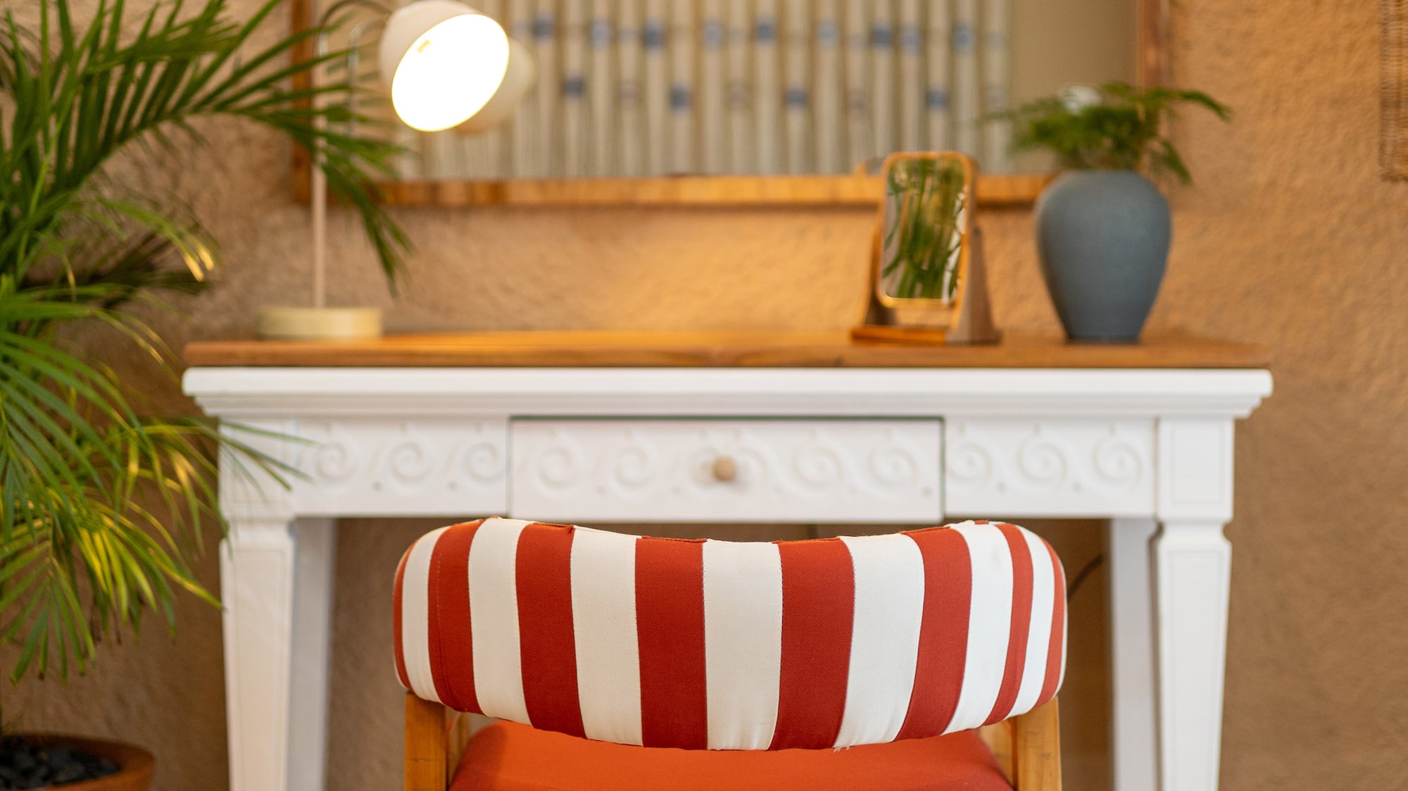 Red striped chair at white vanity table with arched mirror, desk lamp, and potted plant.