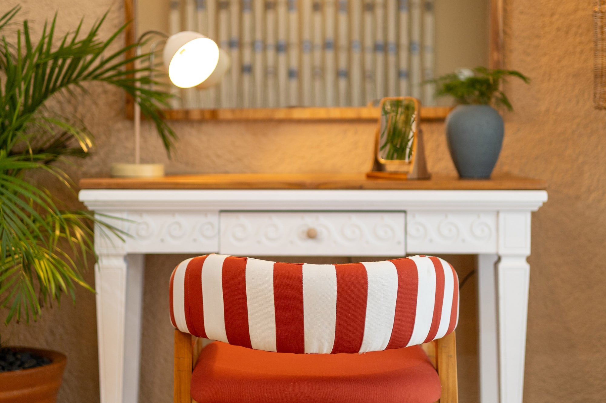 Red striped chair at white vanity table with arched mirror, desk lamp, and potted plant.