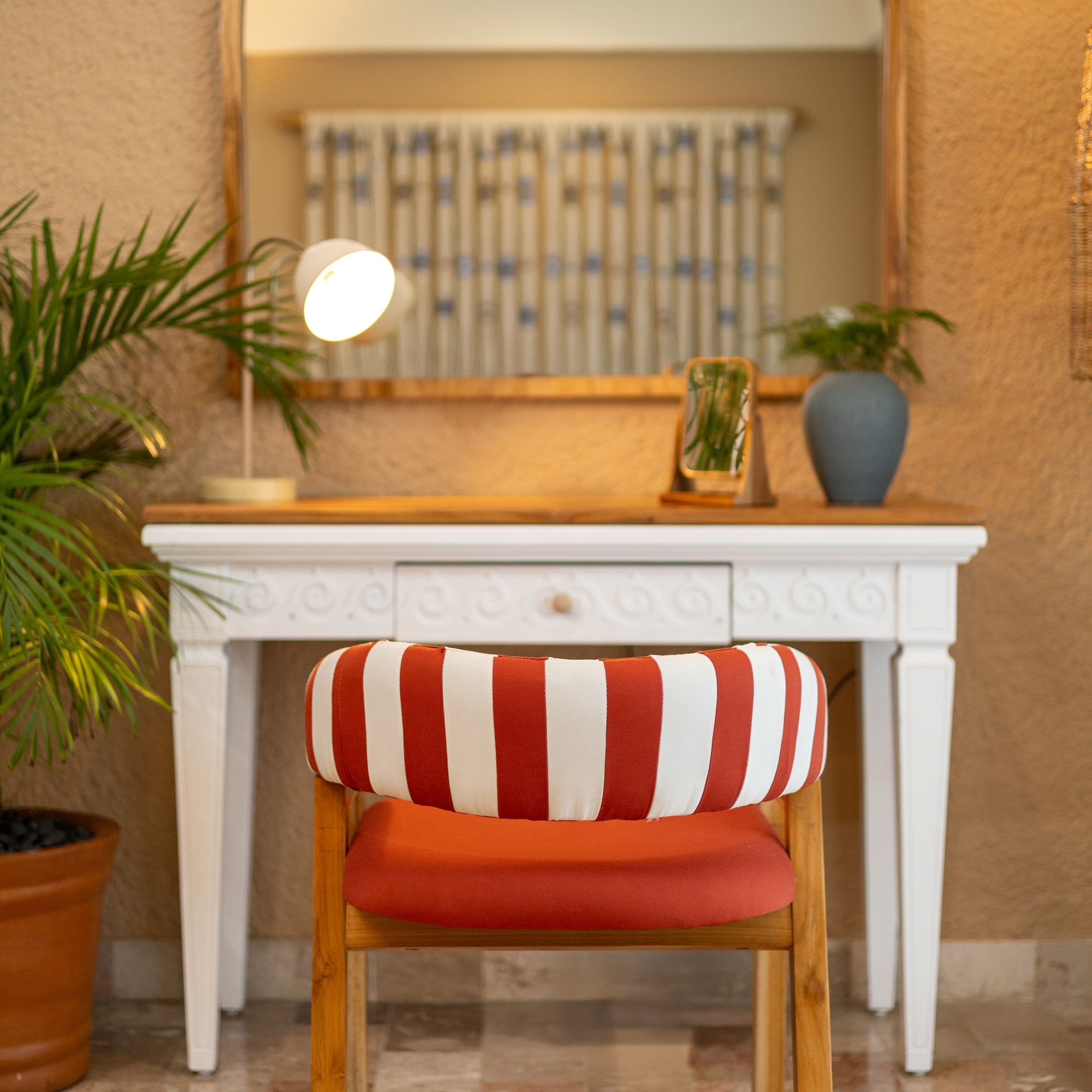 Red striped chair at white vanity table with arched mirror, desk lamp, and potted plant.