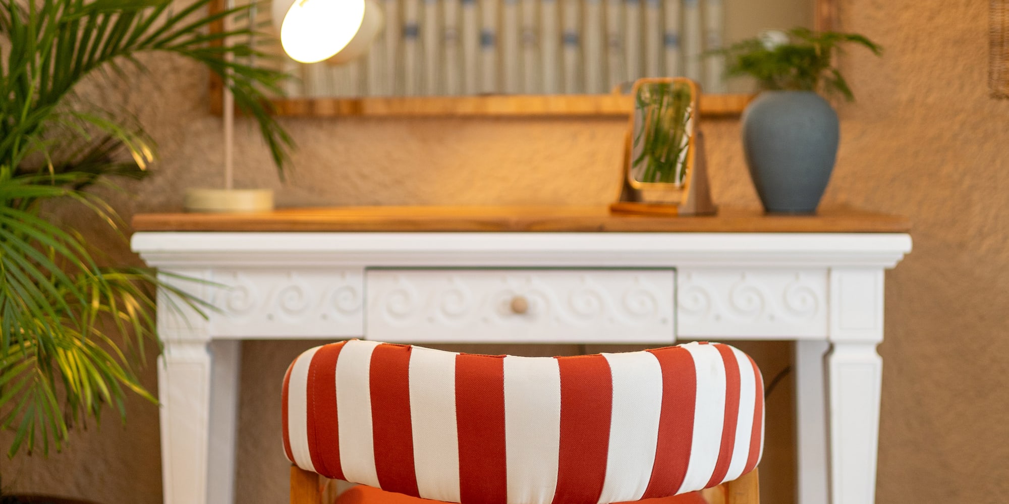 Red striped chair at white vanity table with arched mirror, desk lamp, and potted plant.
