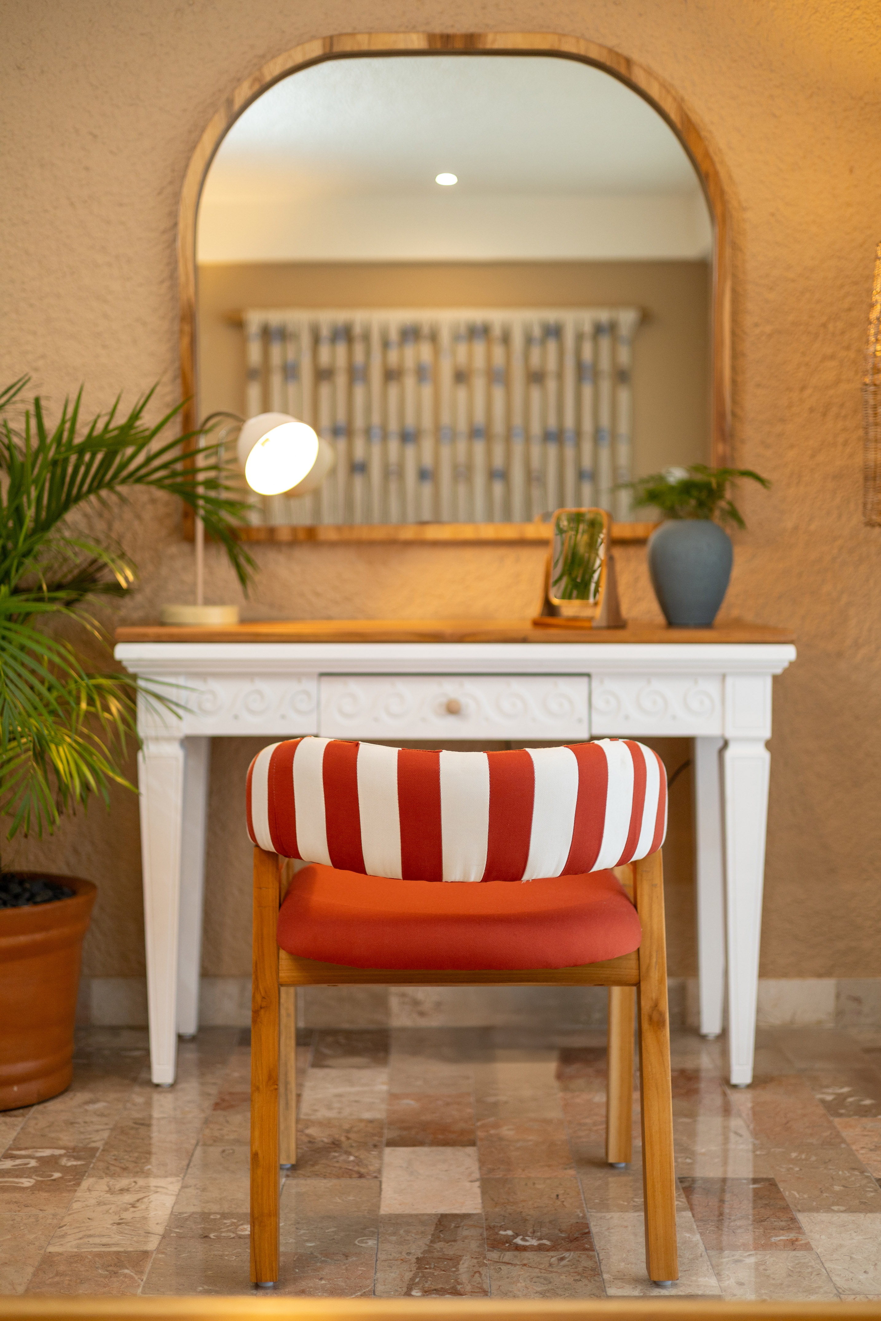 Red striped chair at white vanity table with arched mirror, desk lamp, and potted plant.